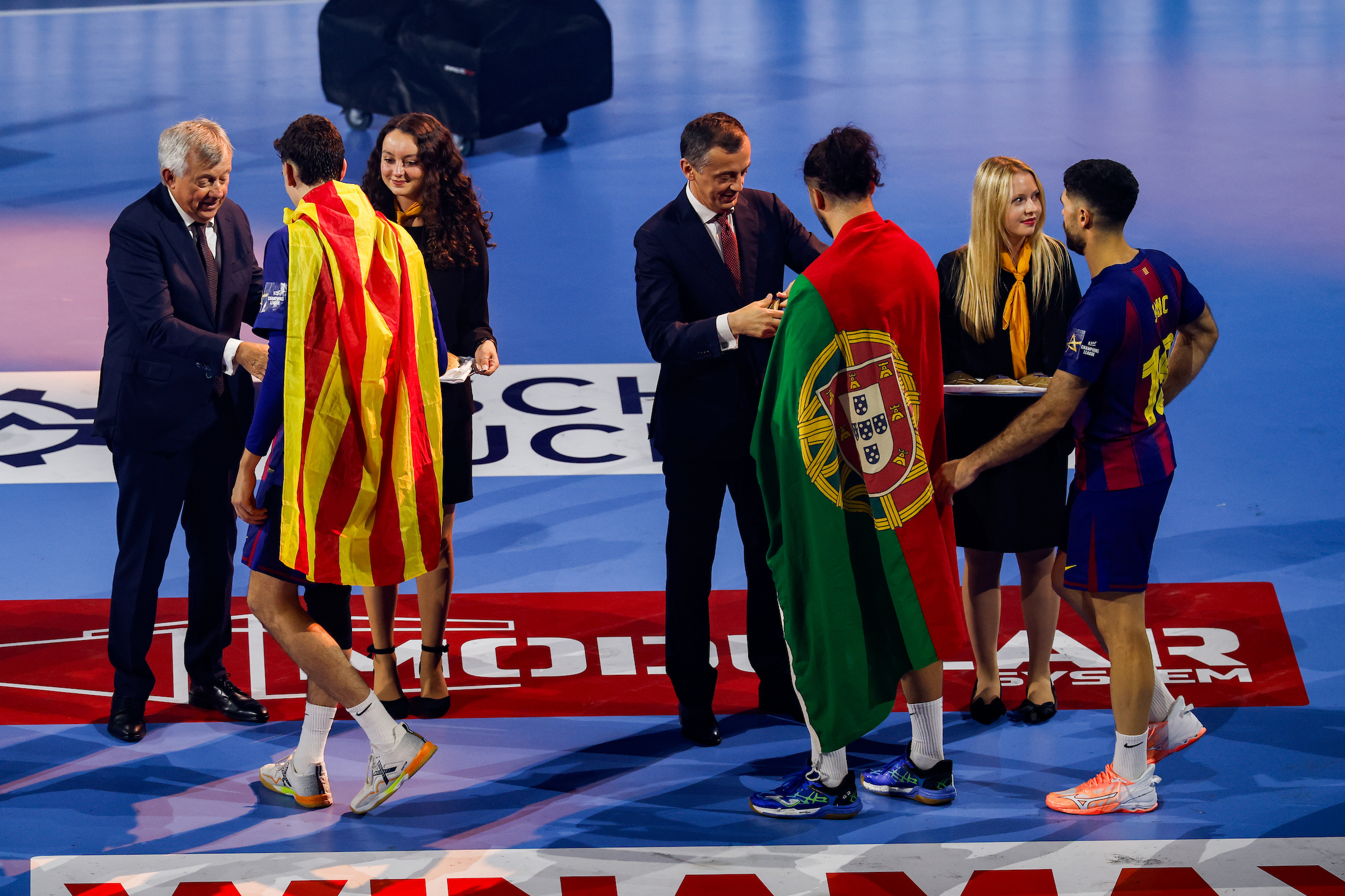 Two athletes draped in national flags stand on a court as officials and staff interact around them.