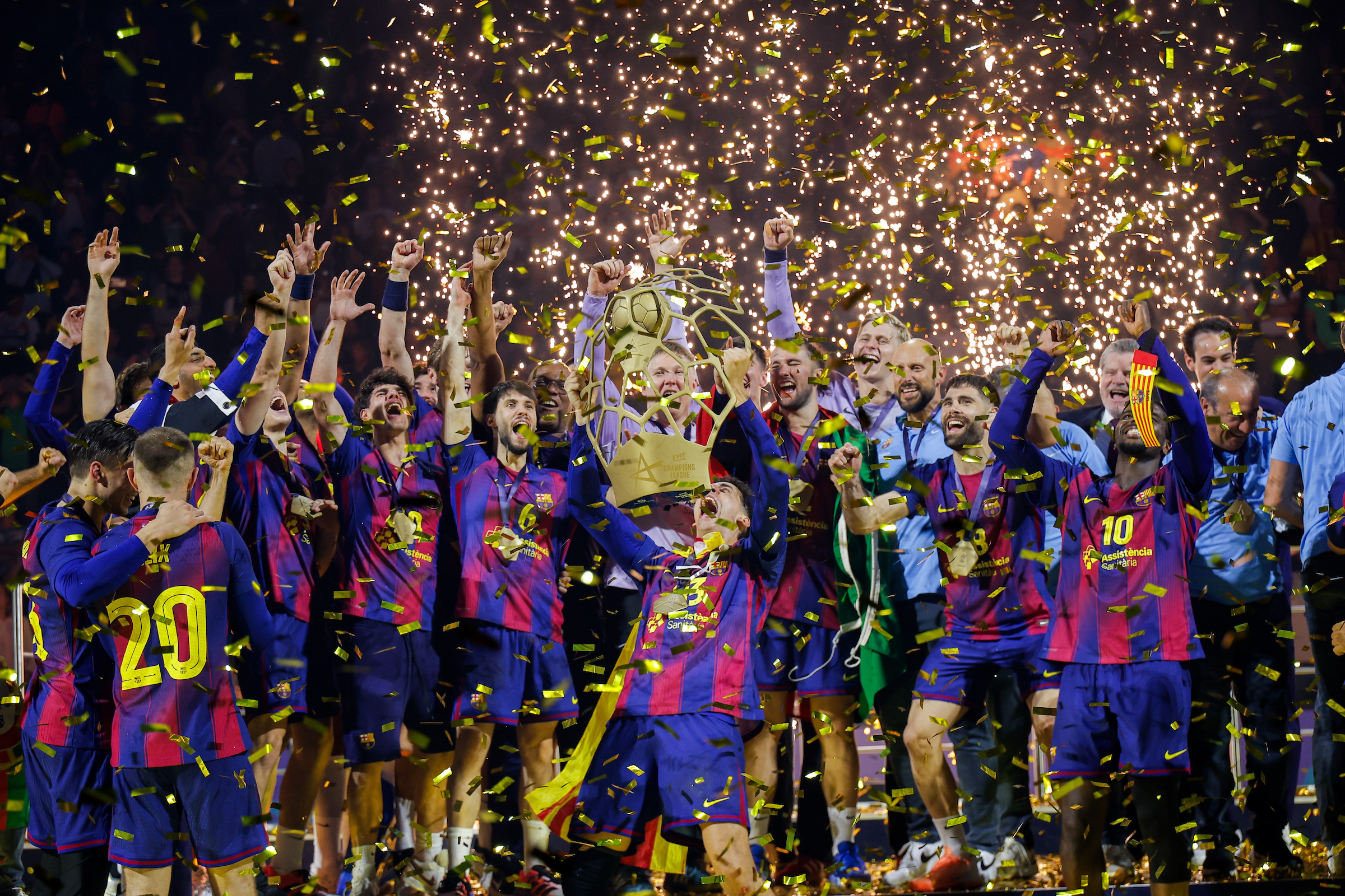 A group of soccer players in blue and red uniforms celebrate with a trophy amidst falling golden confetti and fireworks.