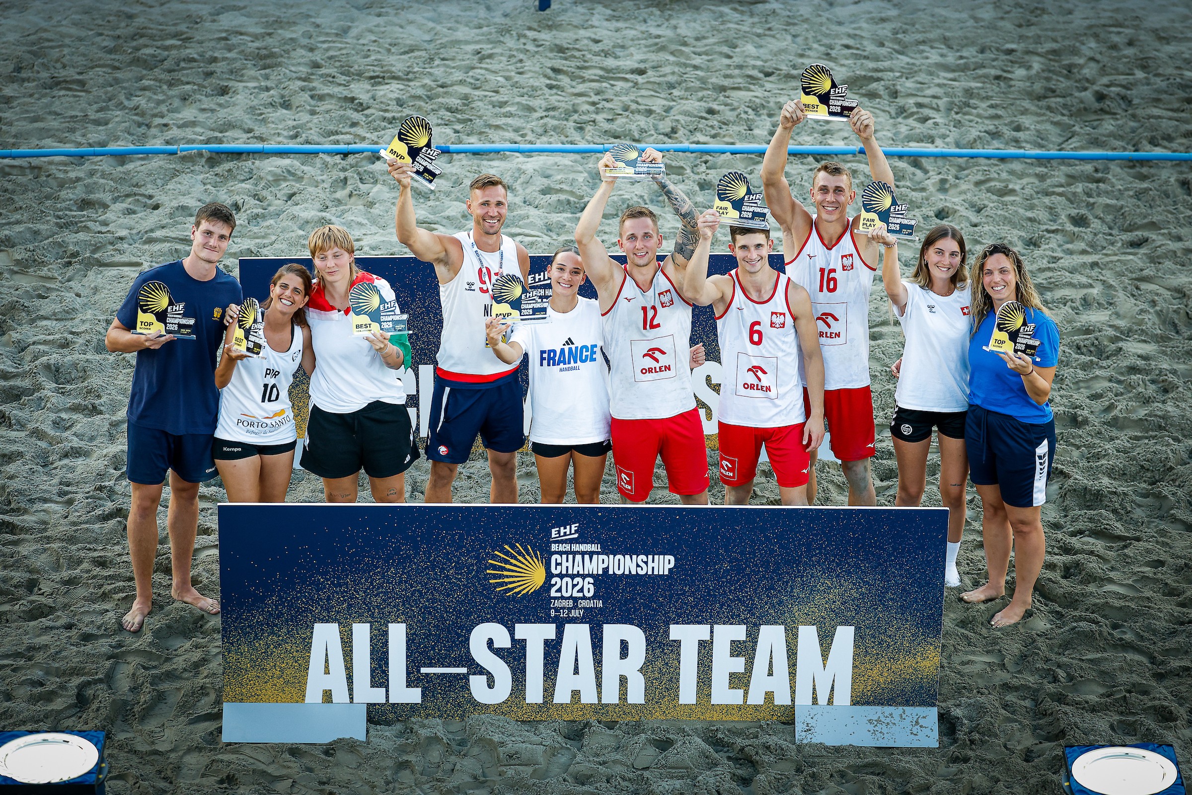 A group of beach volleyball players stands on the sand holding awards, posing behind a sign that reads "All-Star Team" for the EHF Beach Handball Championship 2026.