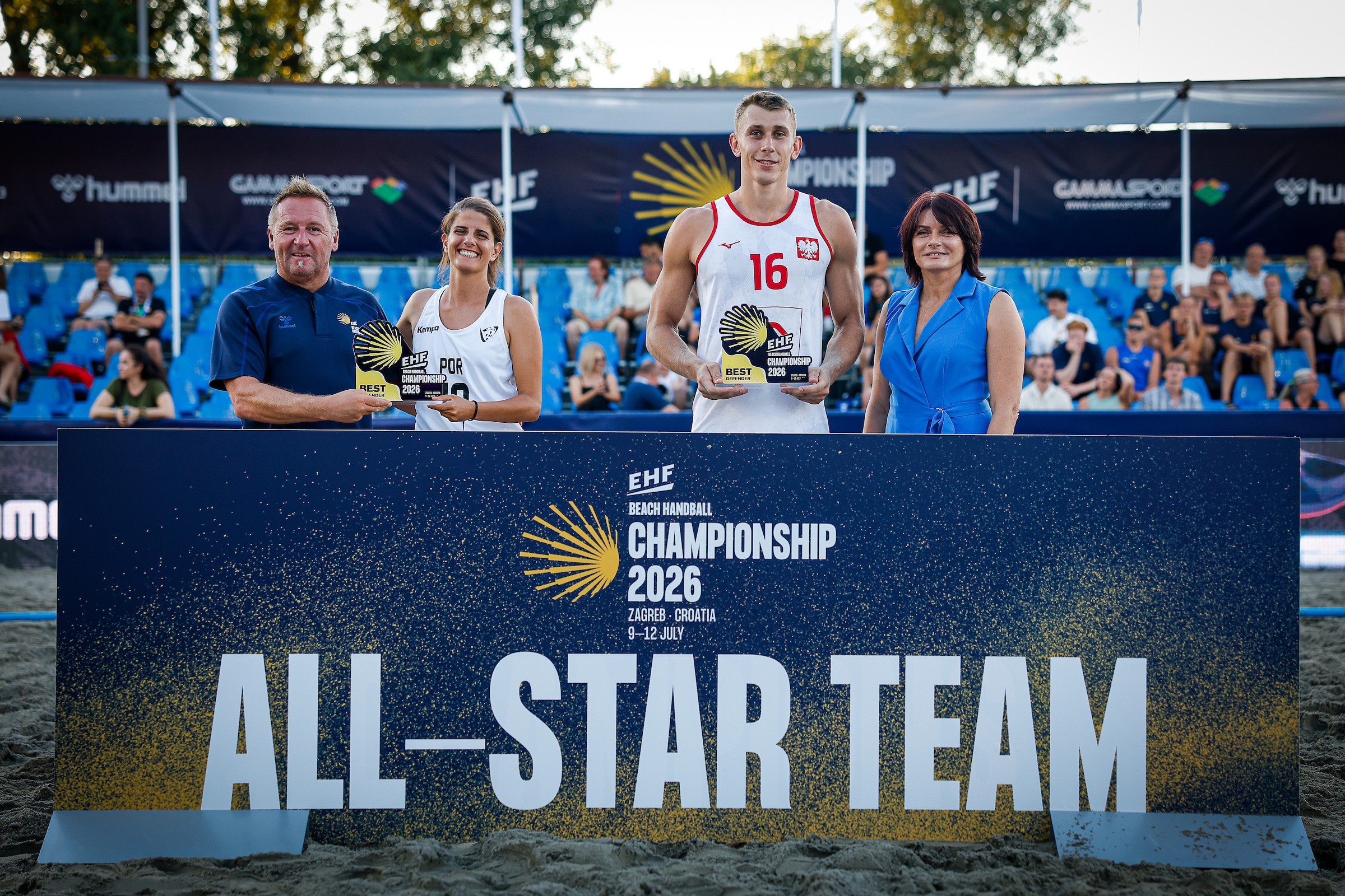 Four individuals stand on a beach volleyball court behind a banner reading “EHF Beach Handball Championship 2026 All-Star Team,” holding awards and smiling for the camera.