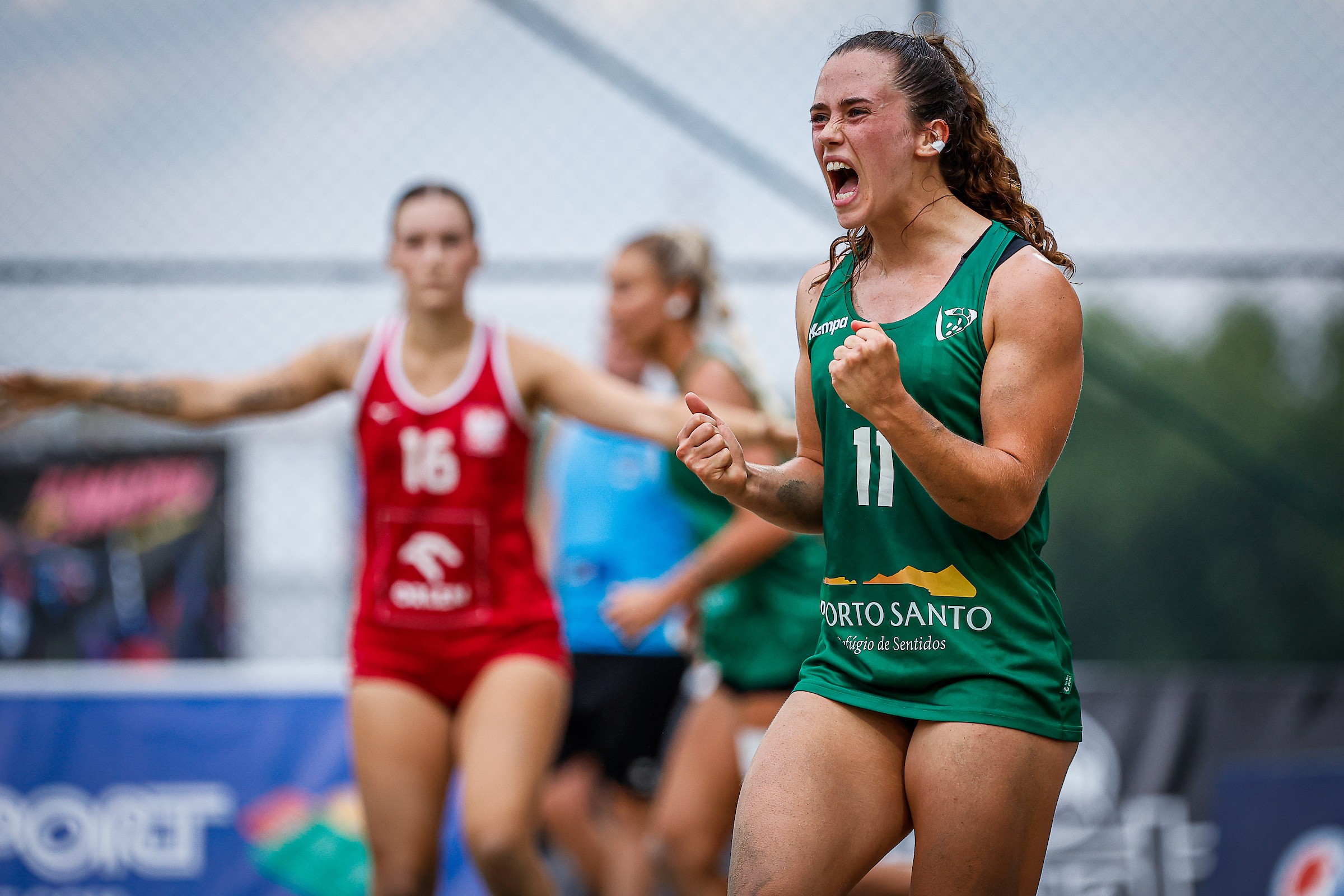 A female athlete in a green jersey celebrates with a raised fist and an open-mouthed yell during a sports match.