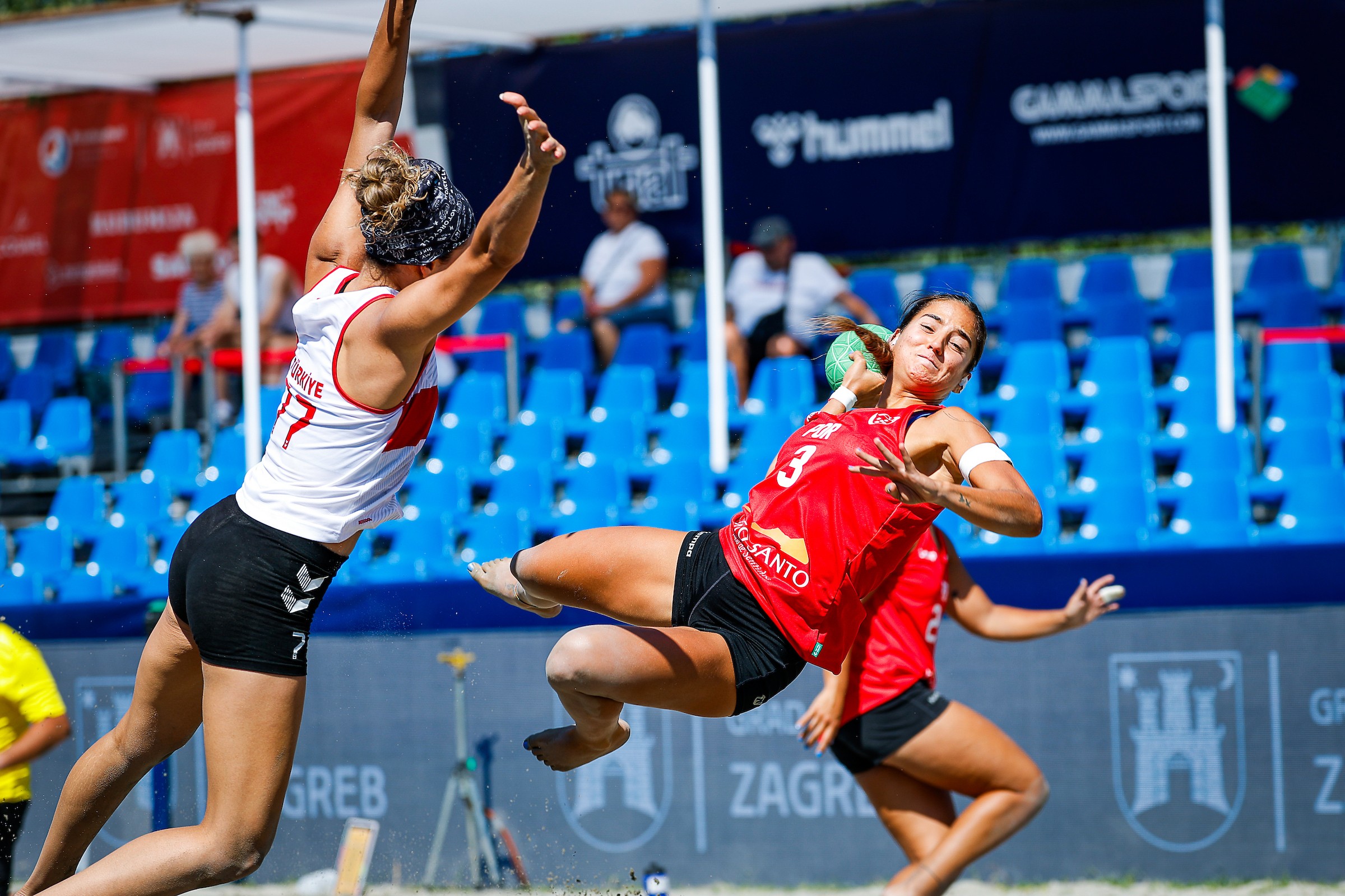 Two athletes compete in a dynamic beach handball match, with one player leaping to block a pass while the other dives to catch the ball.