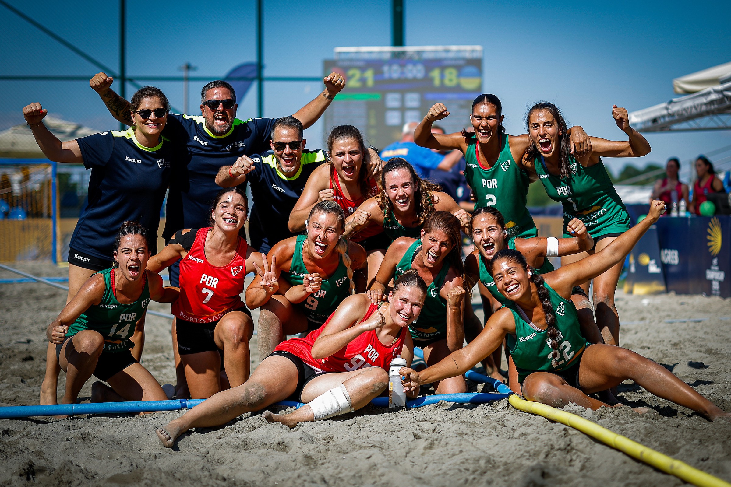 A beach volleyball team in green and red uniforms celebrates enthusiastically on the sand with raised fists and joyful expressions.
