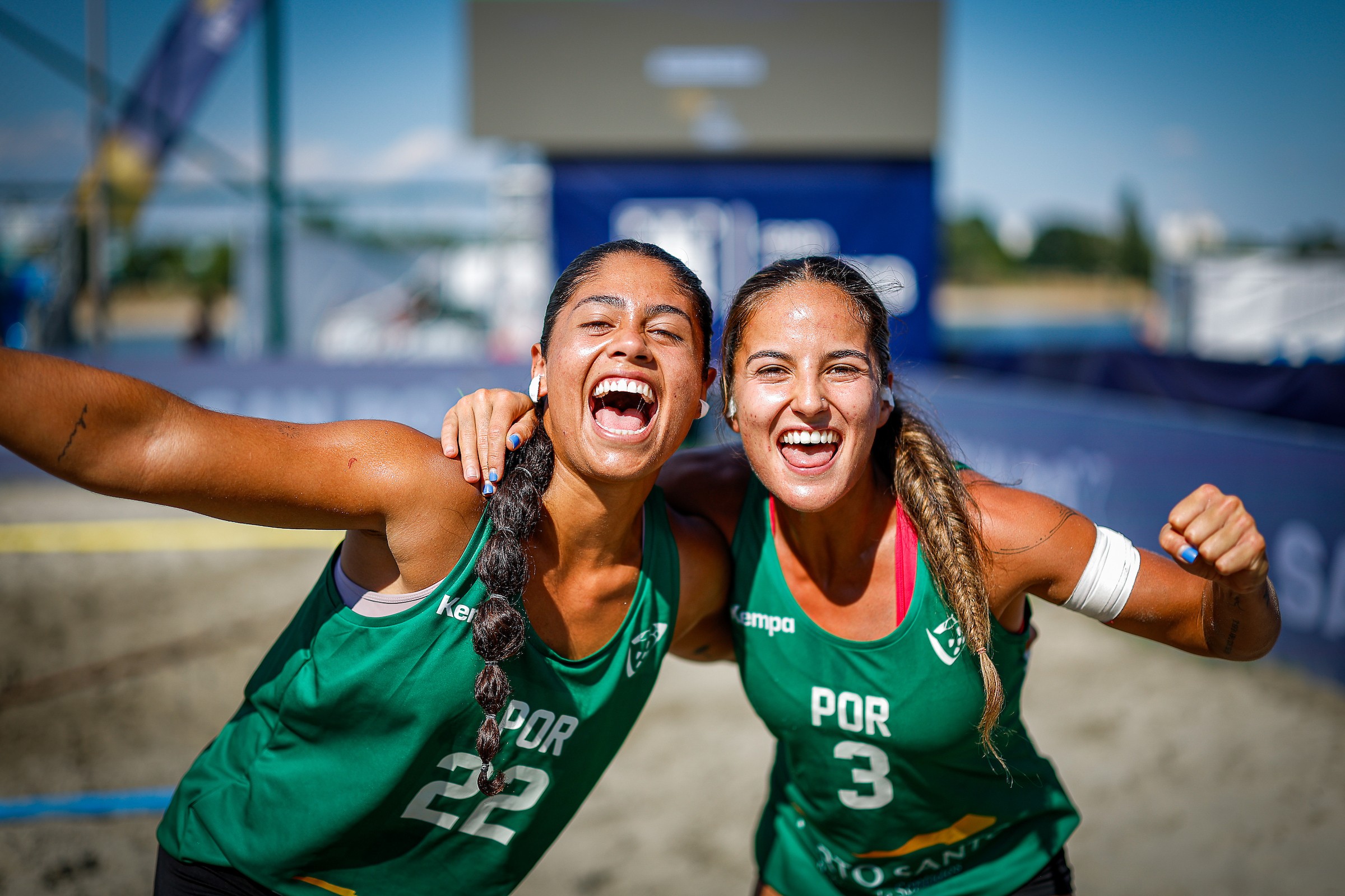 Two athletes in green jerseys celebrate joyfully on a sandy court, arms around each other with wide smiles.