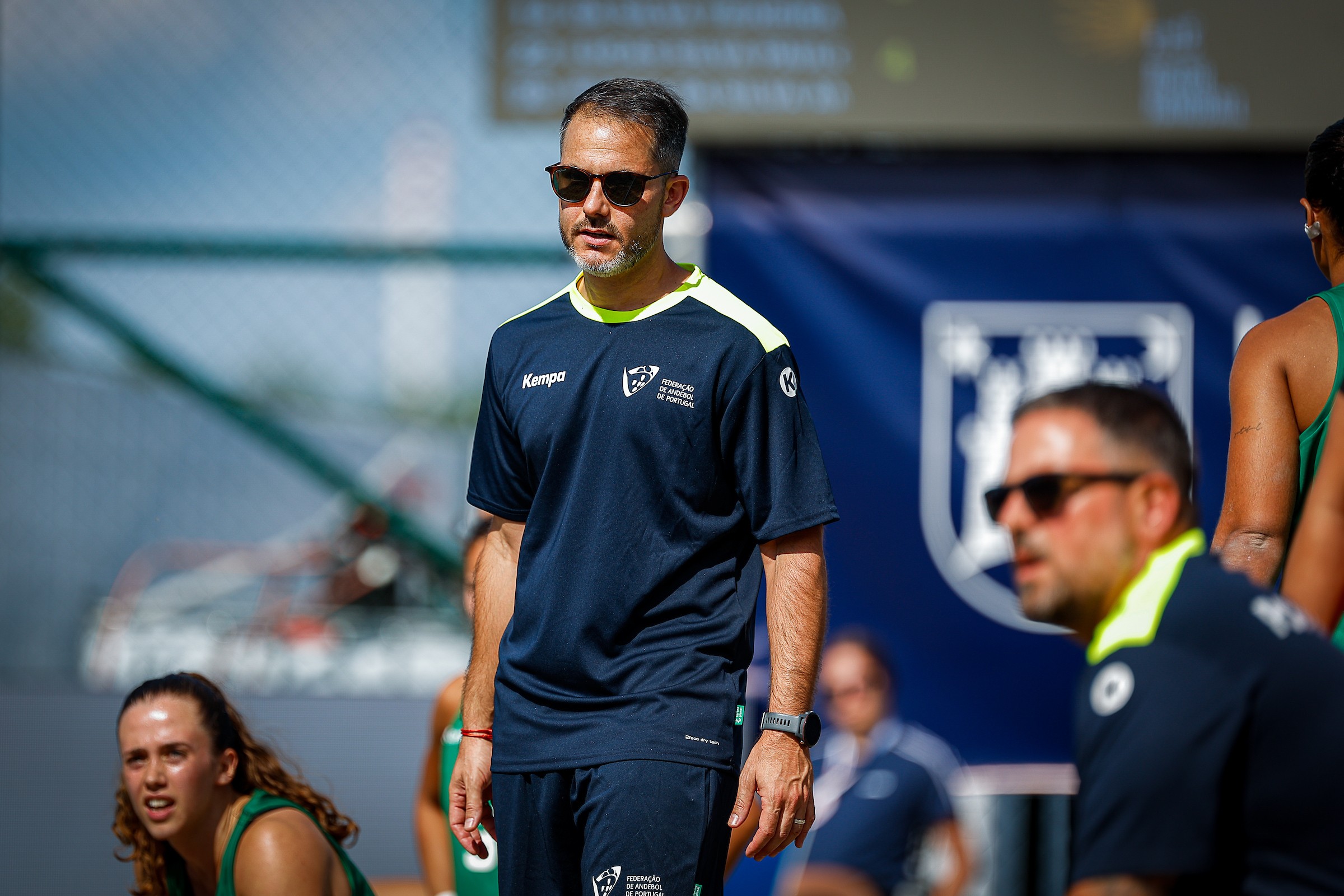 A coach in a navy sports uniform and sunglasses stands on a tennis court, observing players during practice.