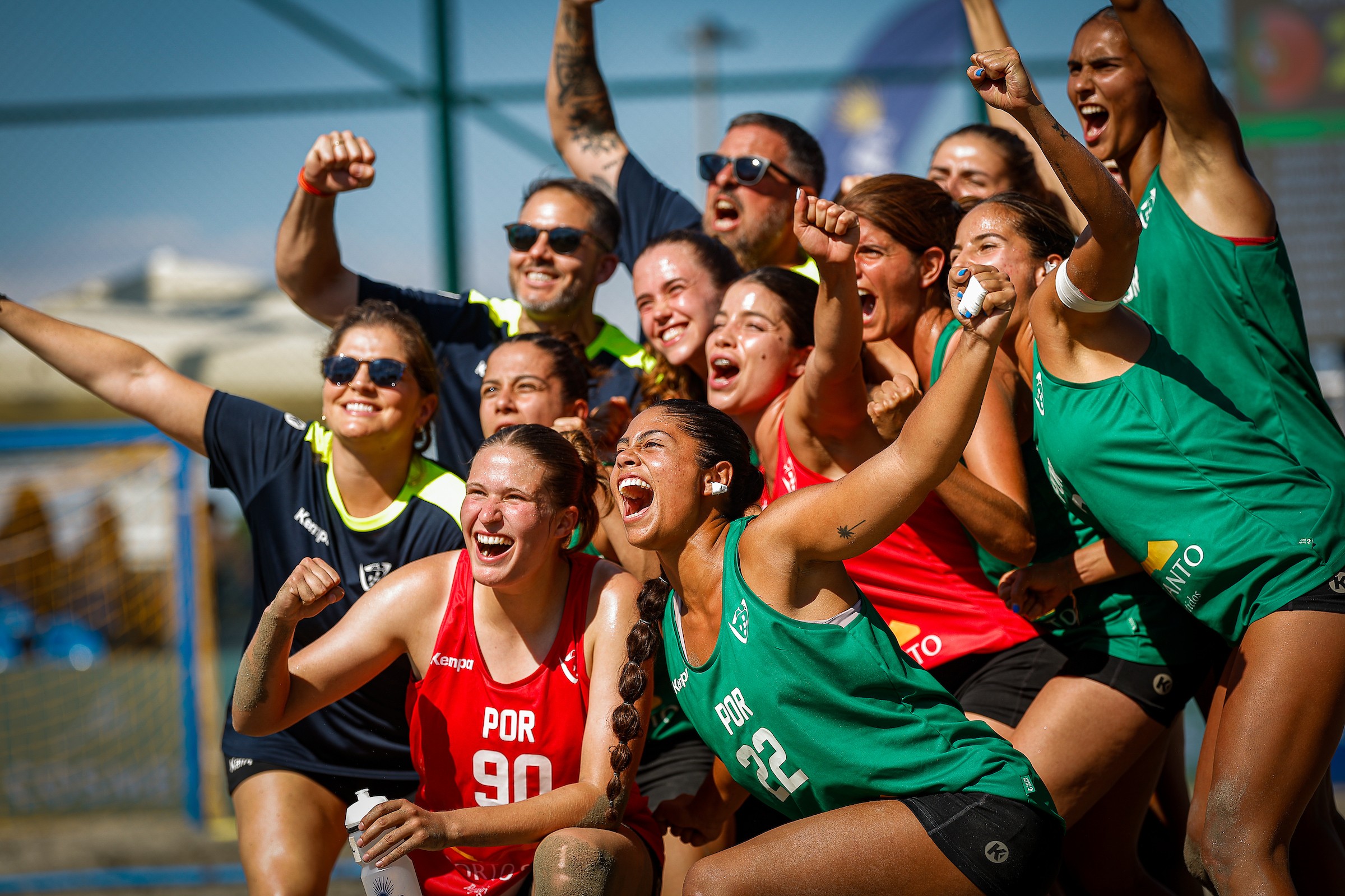A group of athletes in green and red uniforms celebrate enthusiastically with raised fists and joyful expressions on a sports field.