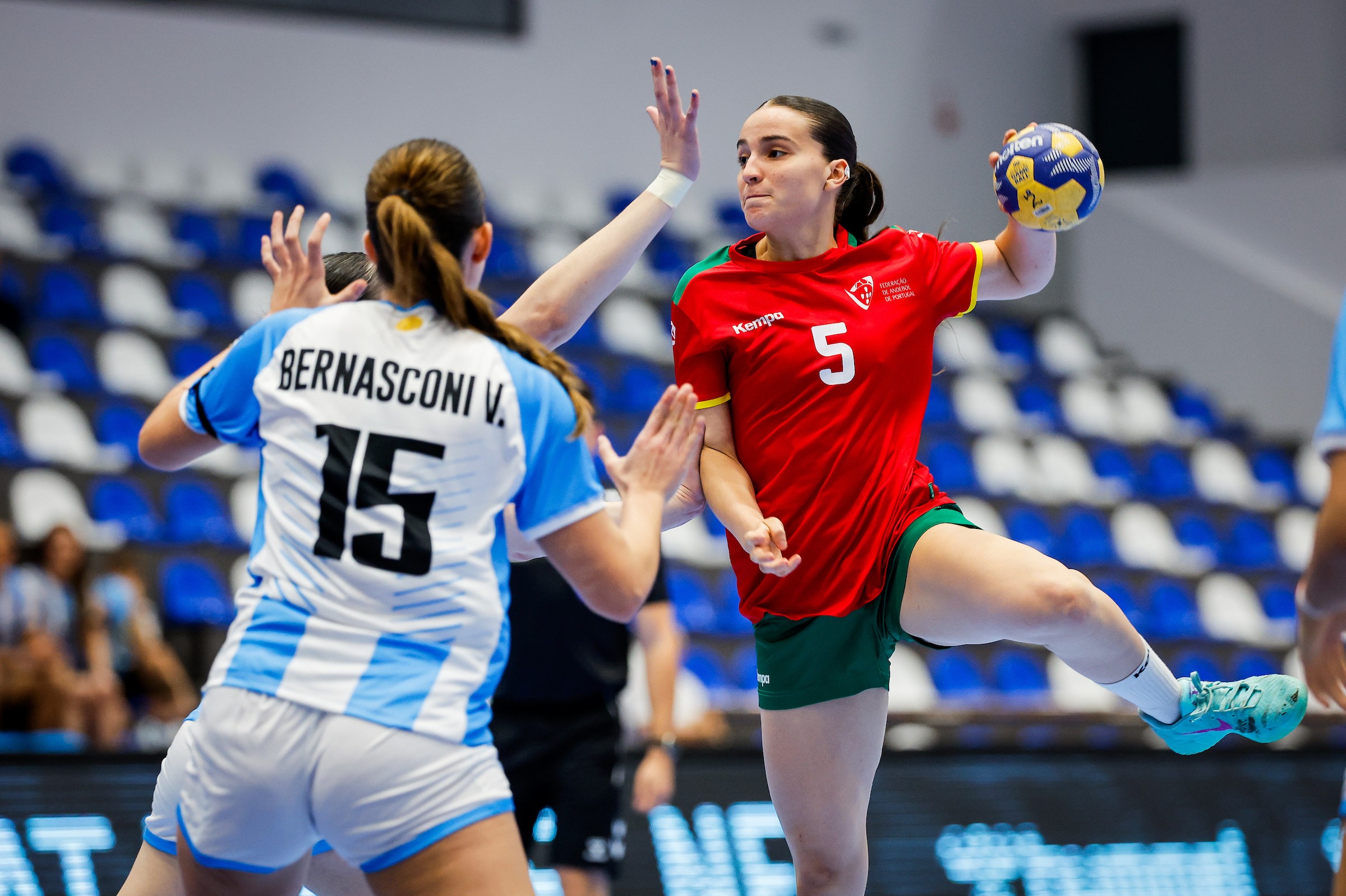 A player in a red jersey with the number 5 leaps to pass a handball while being defended by an opponent in a blue and white jersey.