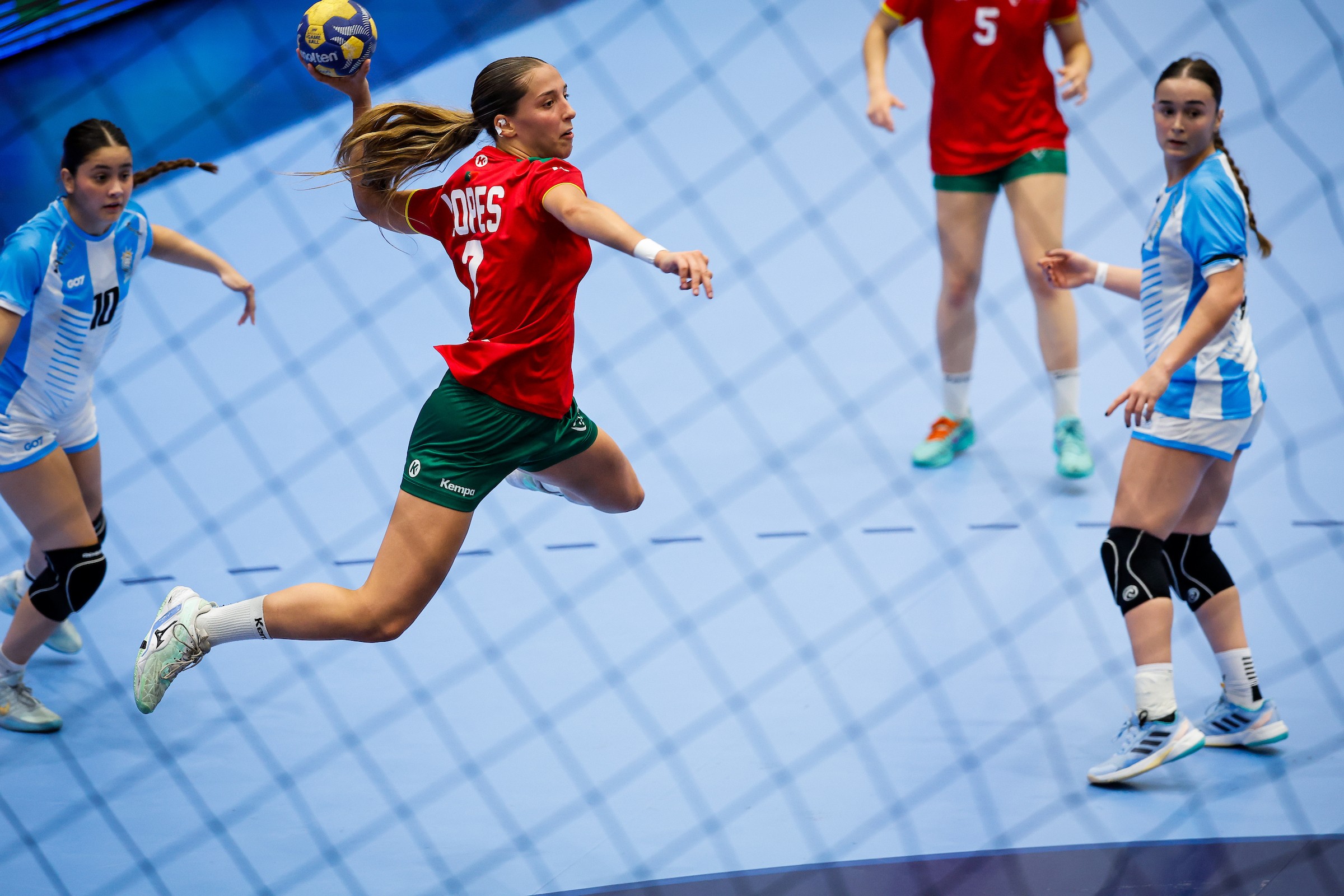 An athlete in a red jersey leaps to pass a handball while opponents in blue and white uniforms defend during a match.