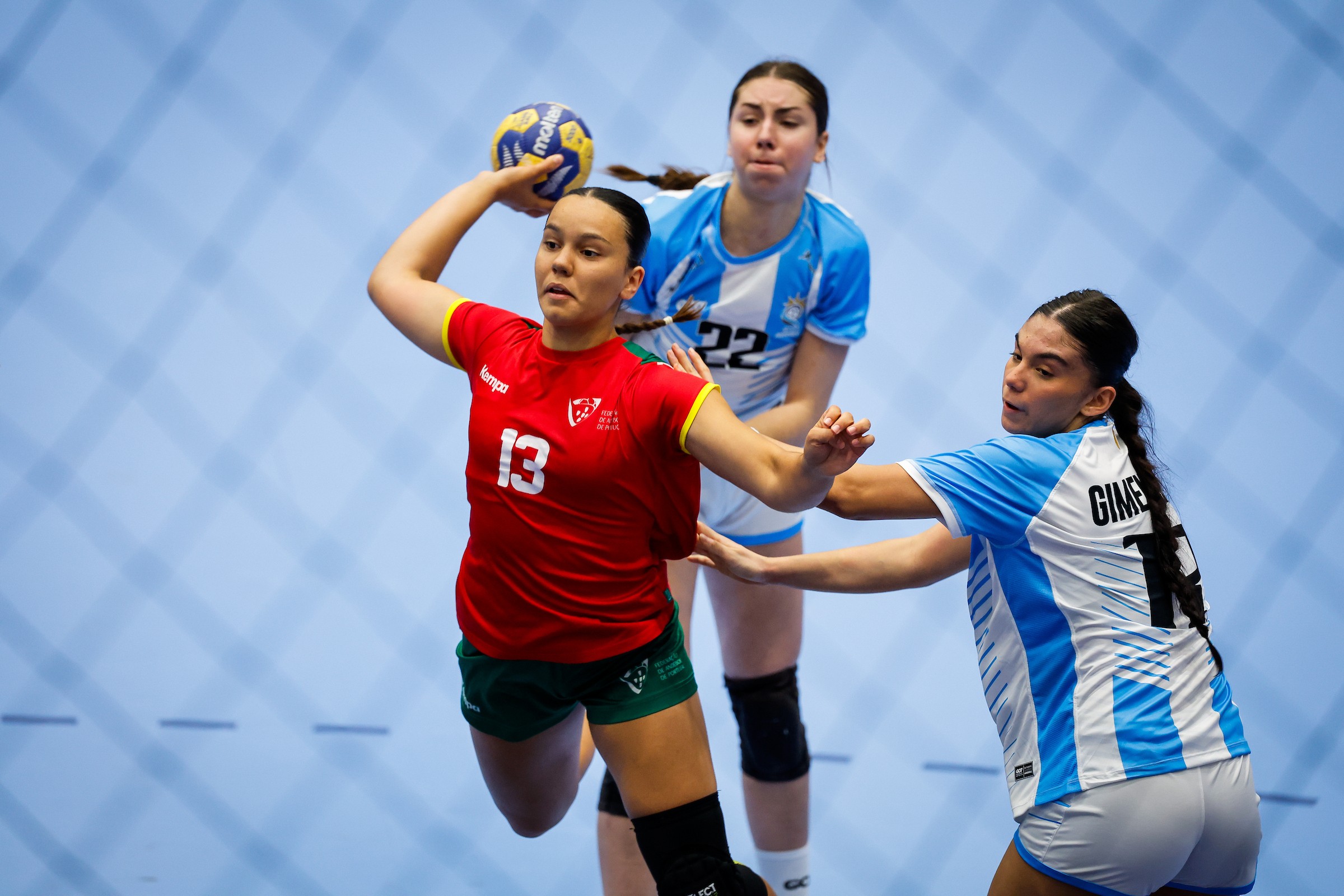 A player in a red jersey prepares to pass the ball while being defended by two opponents in blue and white uniforms during a handball match.