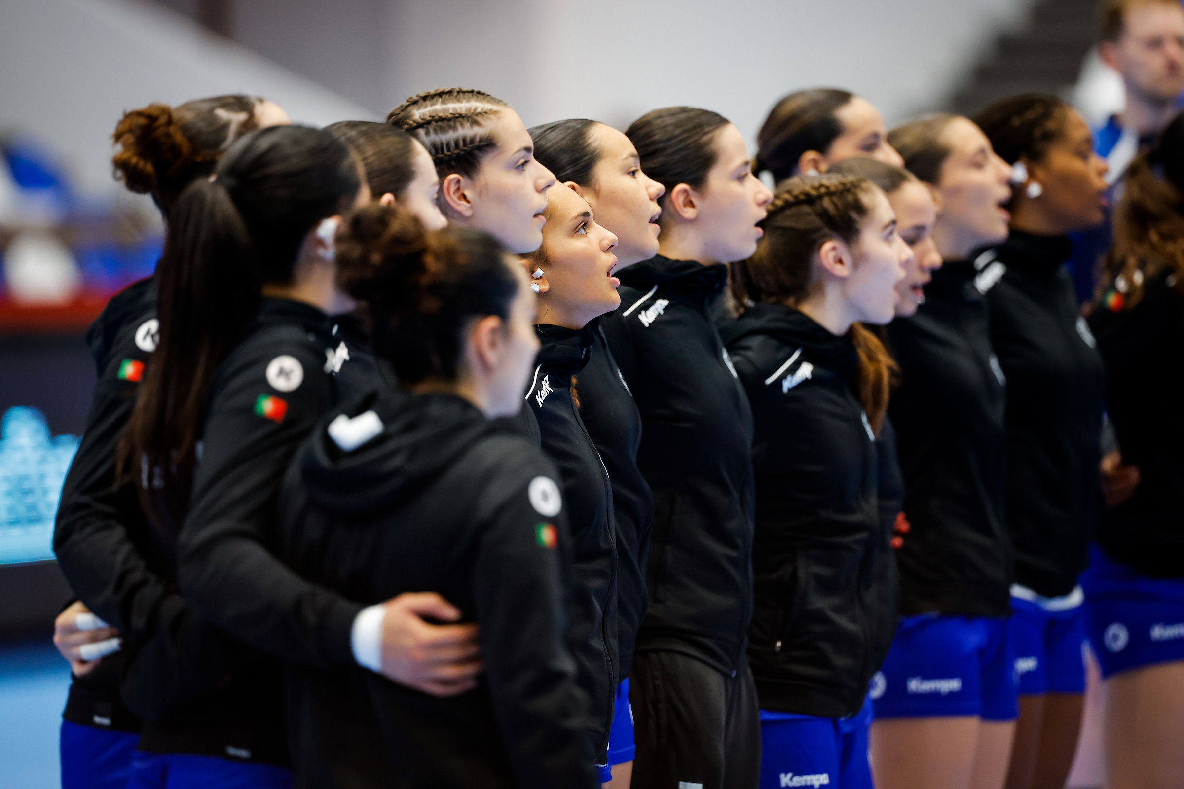 A line of athletes in black jackets and blue shorts stand in unison, singing or chanting with their mouths open.