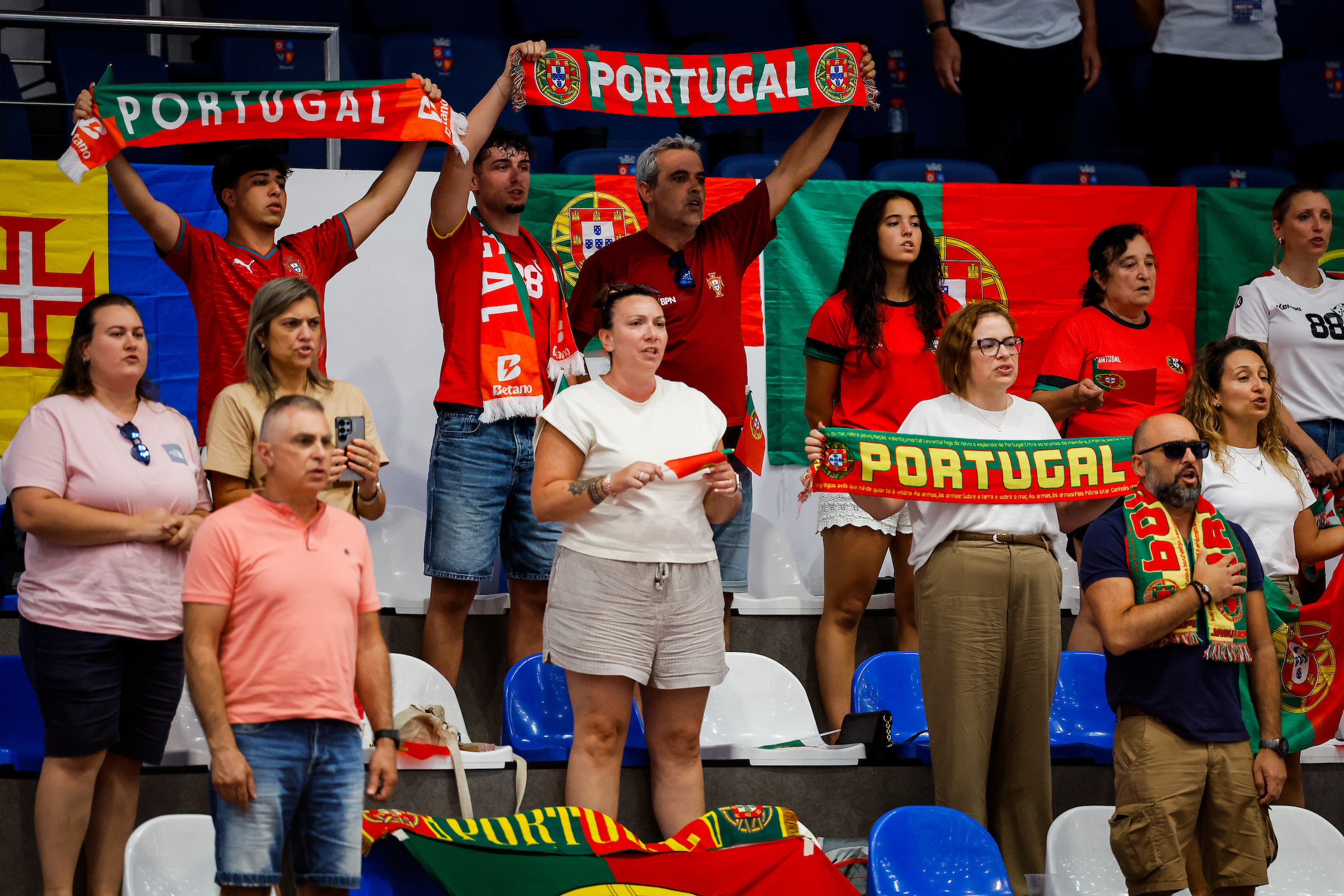 A group of supporters wearing Portugal-themed apparel and holding scarves cheer from stadium seating.