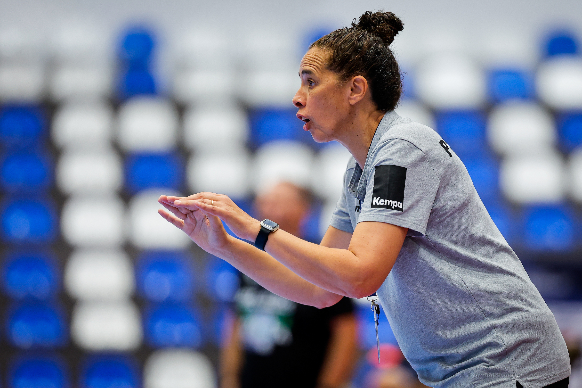 A person in a gray sports shirt with a "Kempa" logo is actively coaching, gesturing with hands extended while speaking during a game.