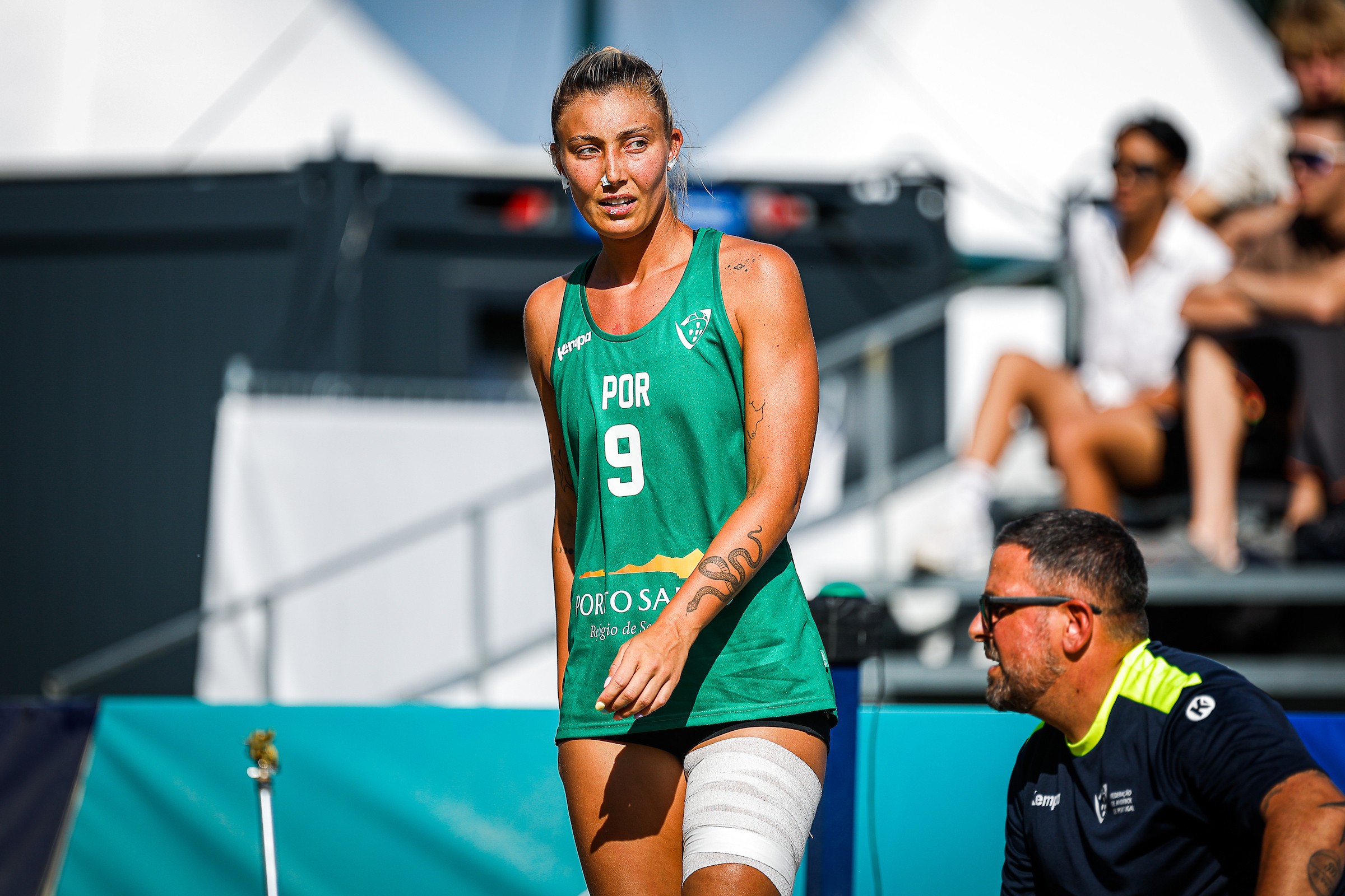 A volleyball player wearing a green jersey with "POR 9" and a bandaged leg stands on the court, while a coach in a dark shirt with neon accents sits nearby.
