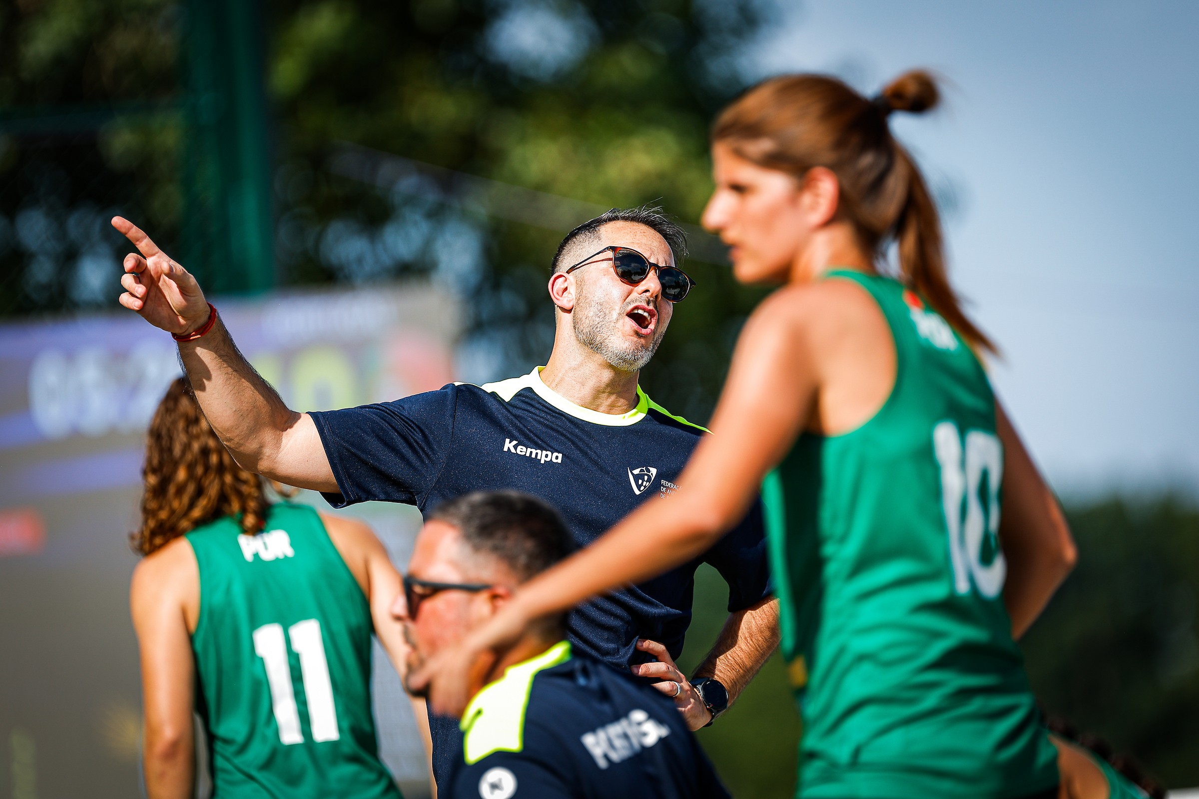 A coach in a navy shirt and sunglasses gestures animatedly while speaking to players in green uniforms during a sports match.