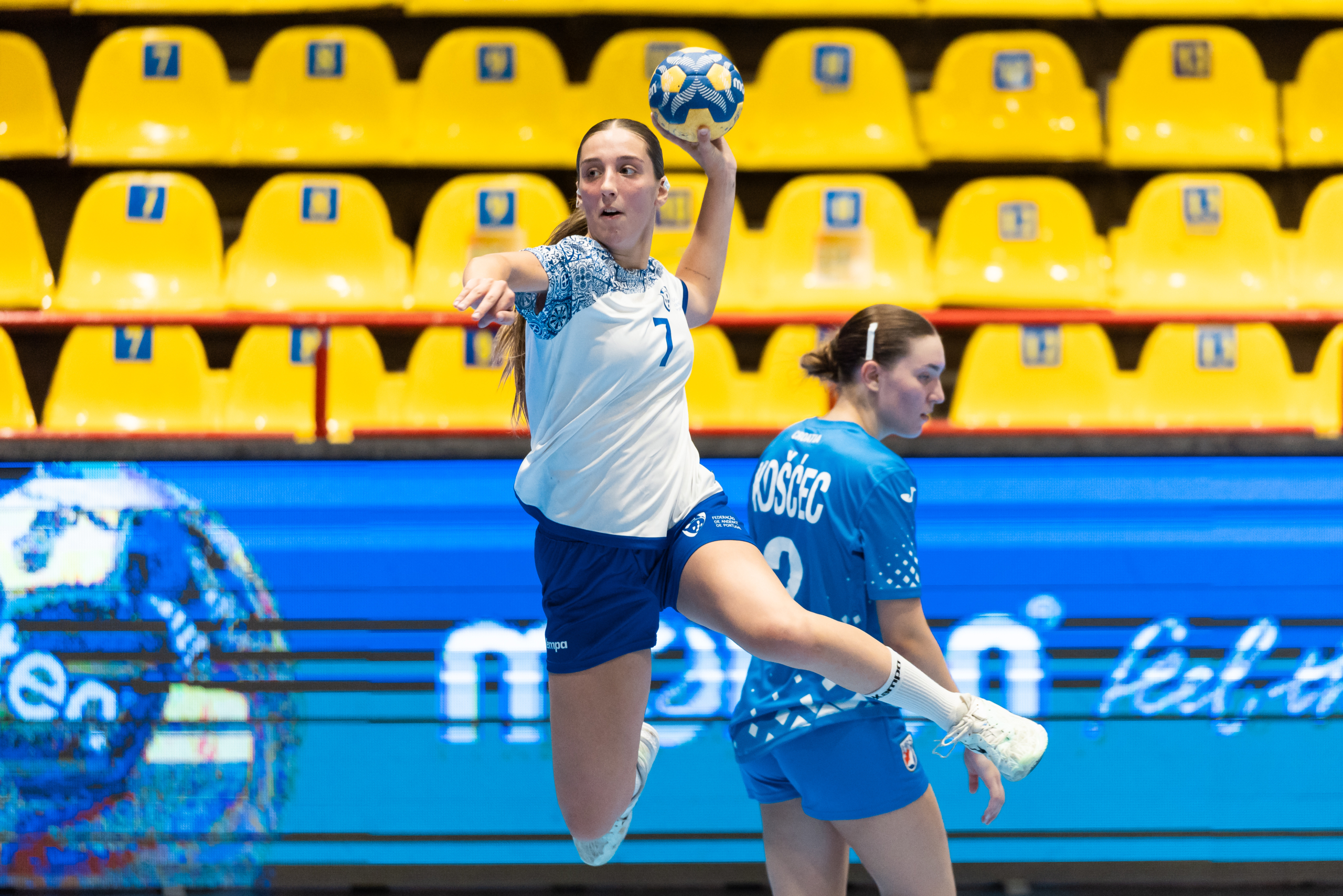An athlete in a white jersey leaps to pass a handball while another player in a blue jersey watches closely, set against yellow stadium seats and a digital display.