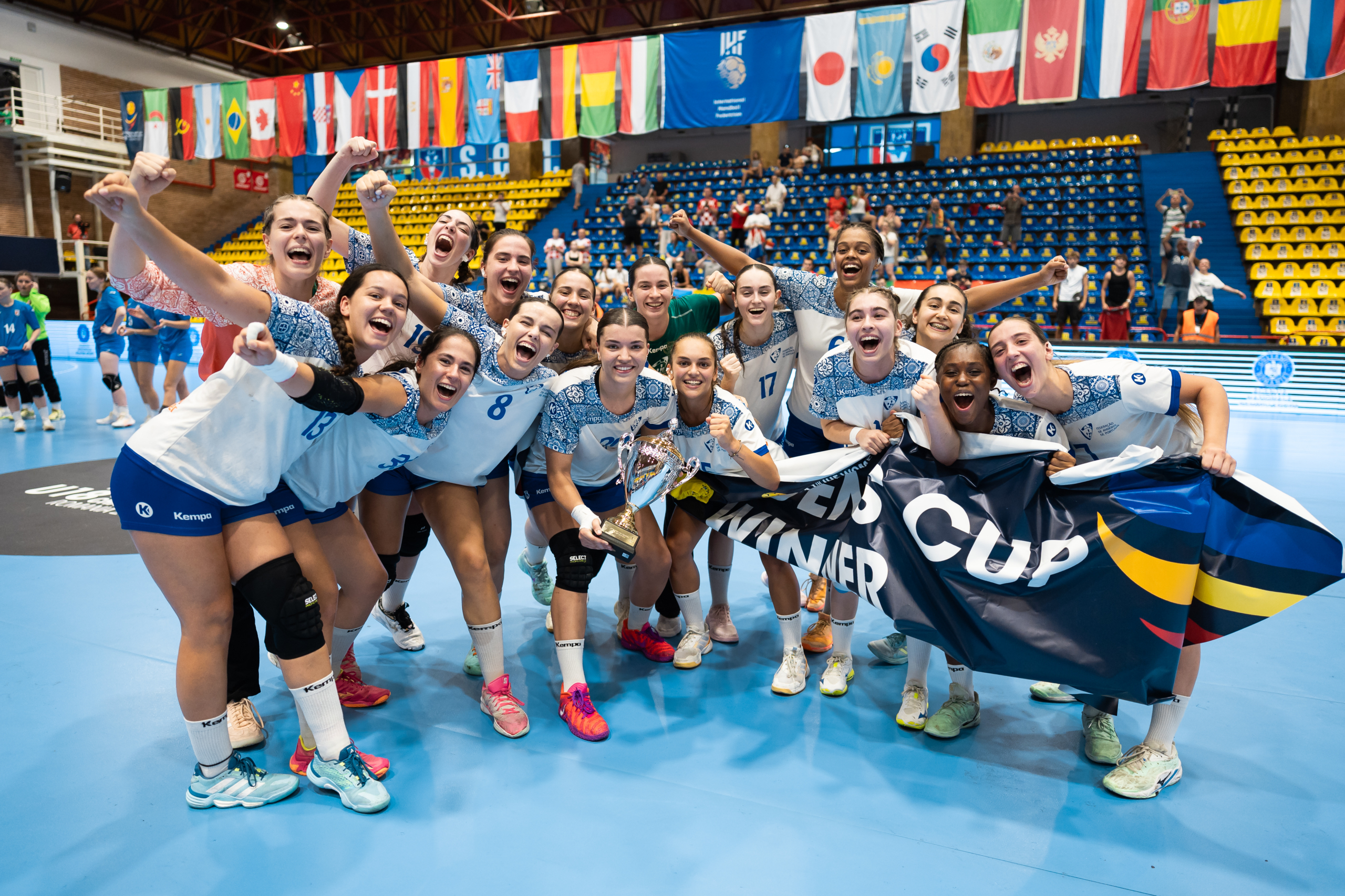 A group of athletes celebrate on a court, holding a trophy and a banner that reads "WINNER," with international flags displayed in the background.