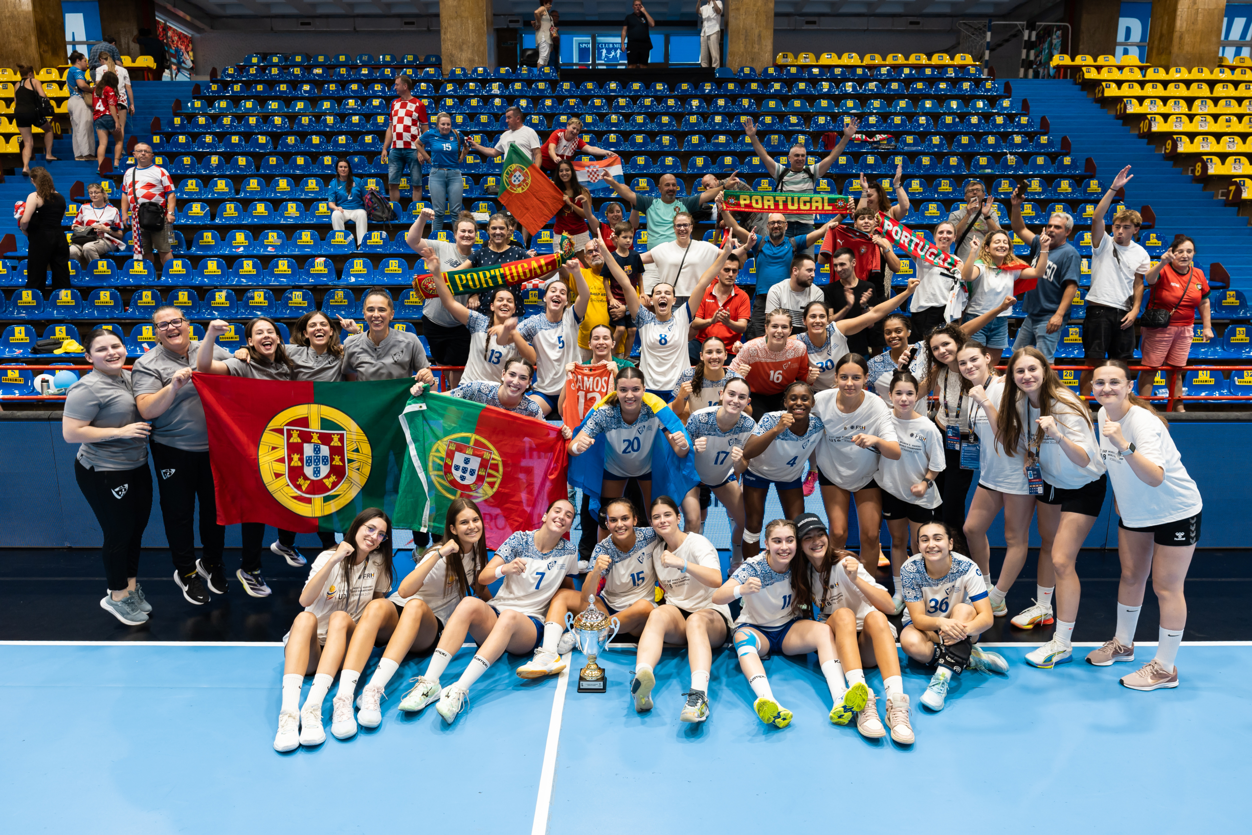 A group of athletes and supporters celebrate with flags and a trophy on a volleyball court.