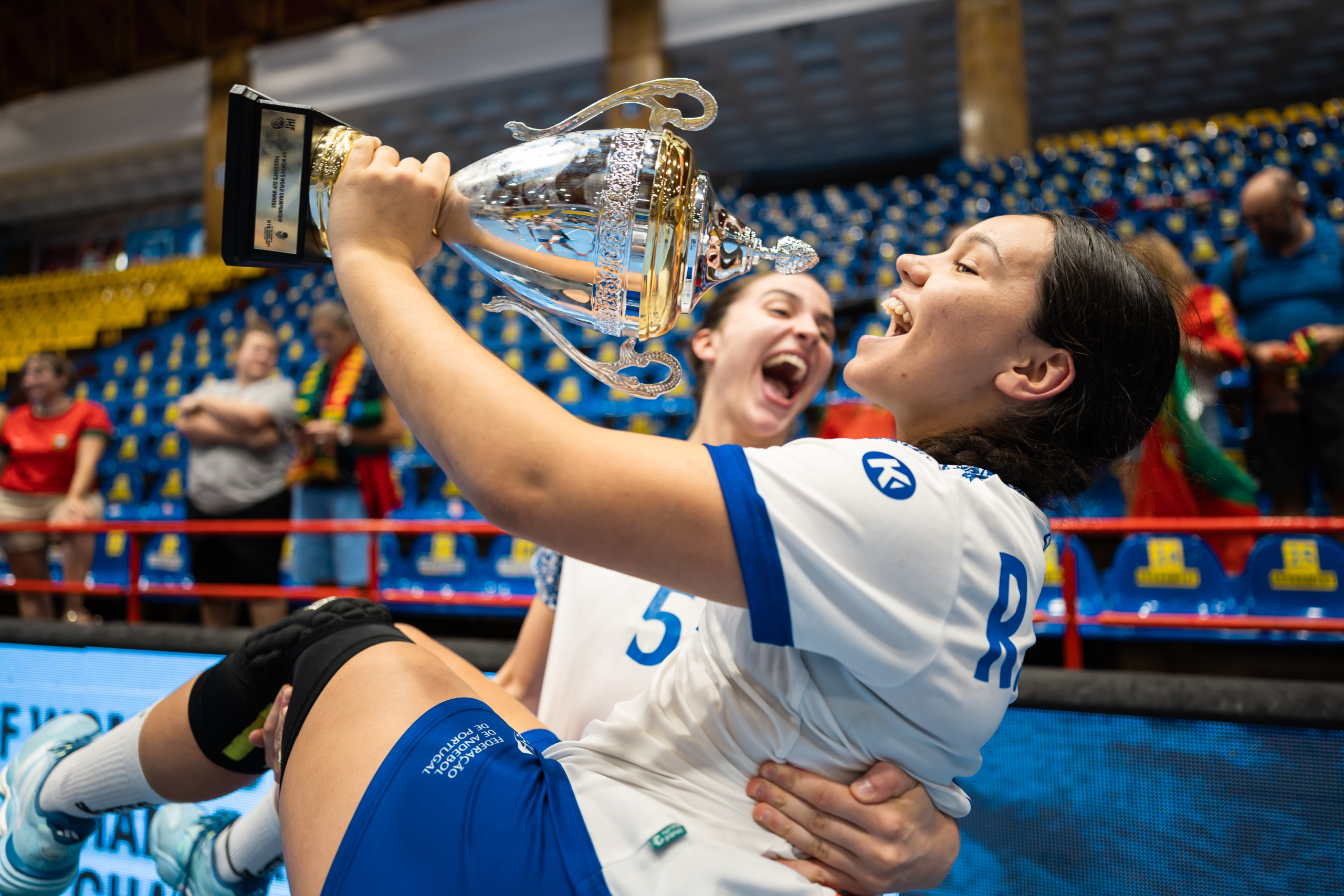 An athlete in a white and blue uniform joyfully lifts a large silver trophy above their head while being embraced by a teammate, with spectators visible in the background.