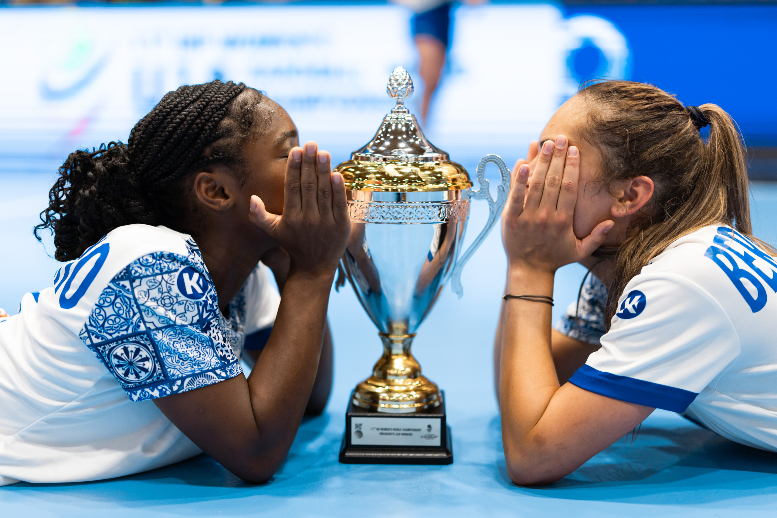Two athletes lean over a trophy, one whispering to the other who covers her face with her hands.