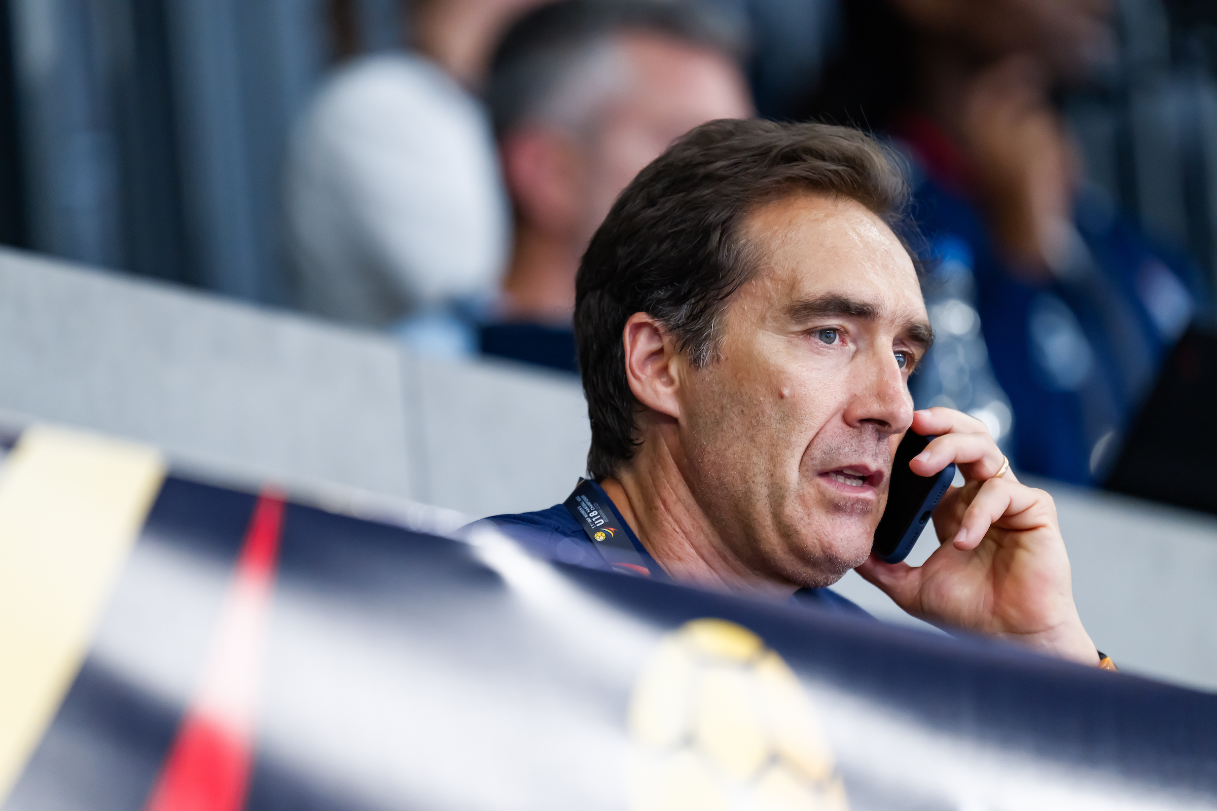 A person with dark hair is speaking on a phone while seated in a stadium, with blurred spectators and a banner in the foreground.