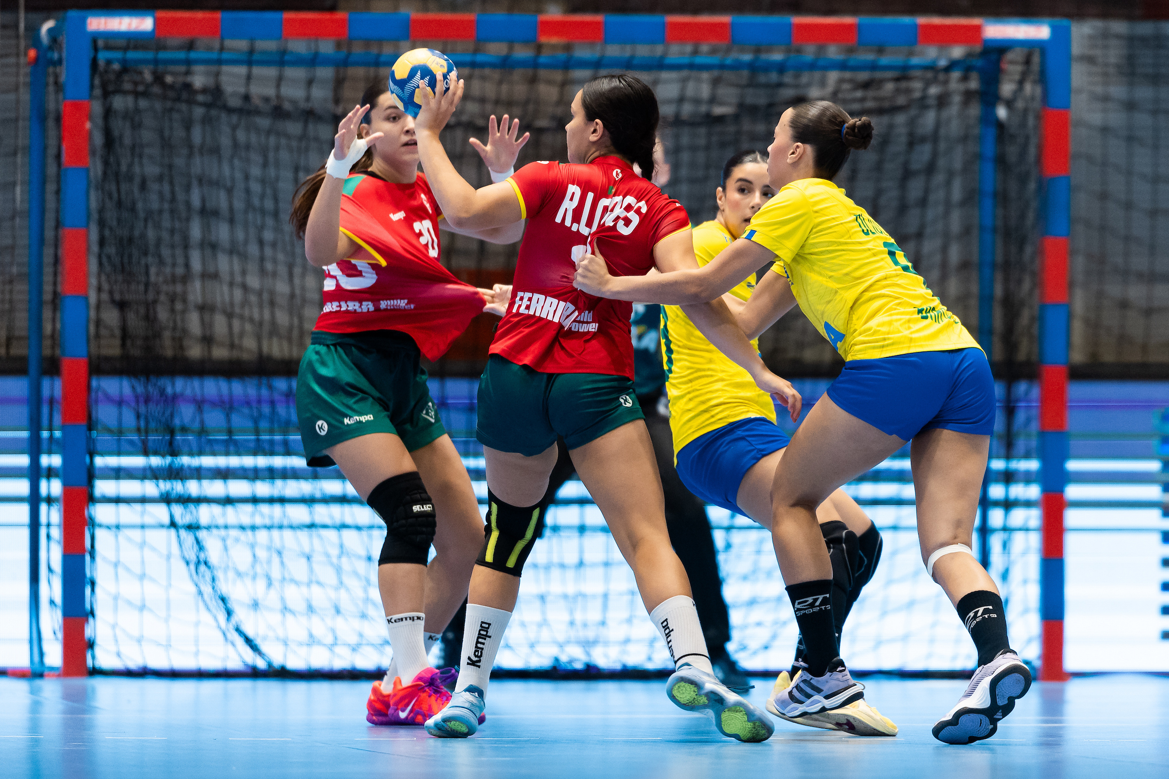 Players in red and yellow jerseys compete for a handball during an indoor match, with one player leaping to catch the ball while others defend and attack.
