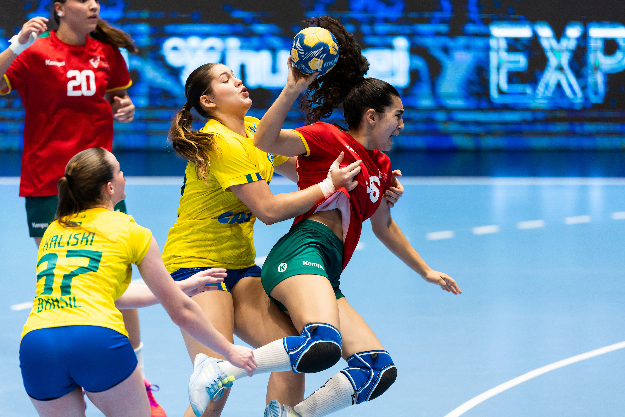 Athletes in yellow and red jerseys compete intensely for a handball during a match on a blue court.