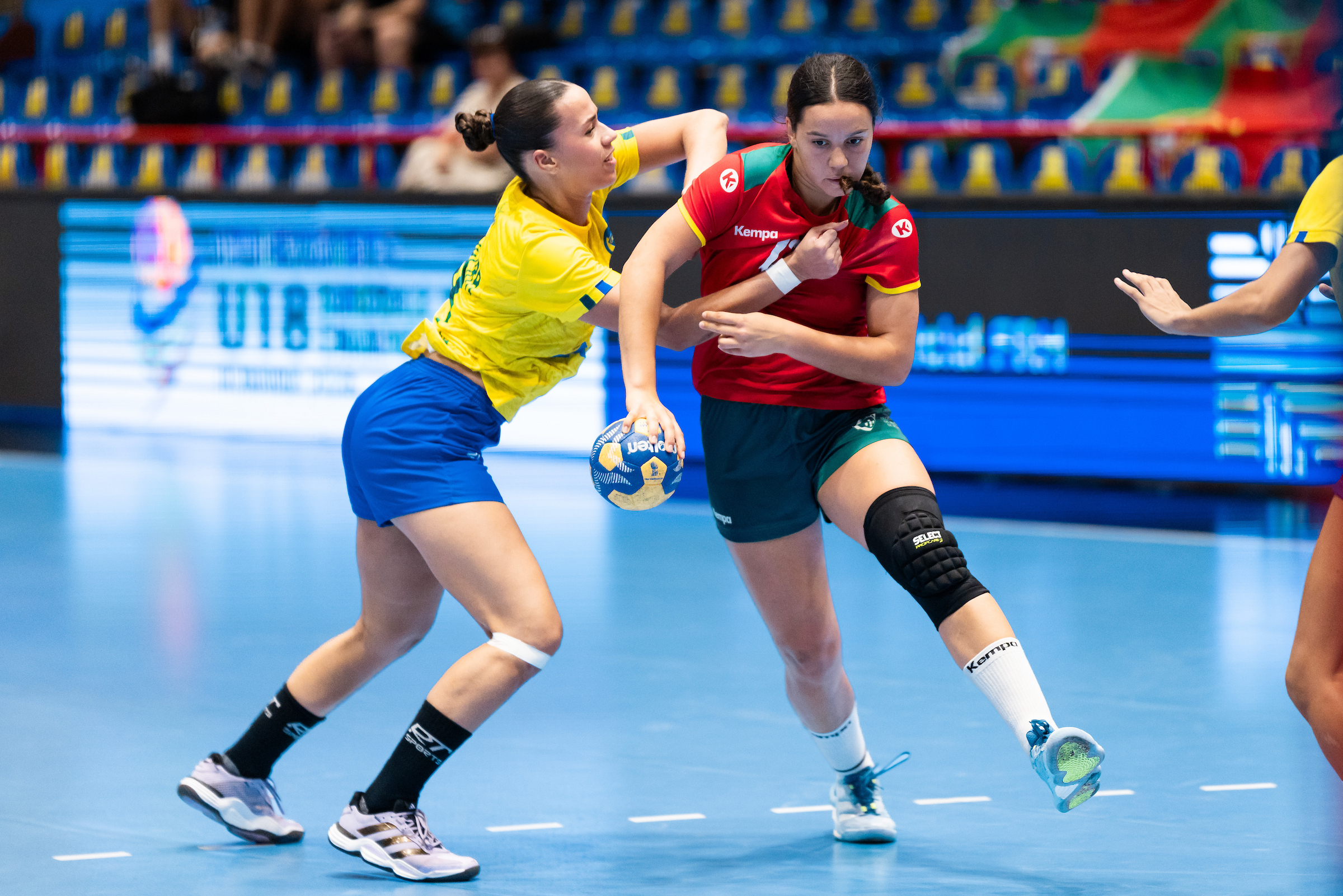 Two athletes compete intensely in a handball match, with one in yellow and blue defending against the other in red and green who is holding the ball.