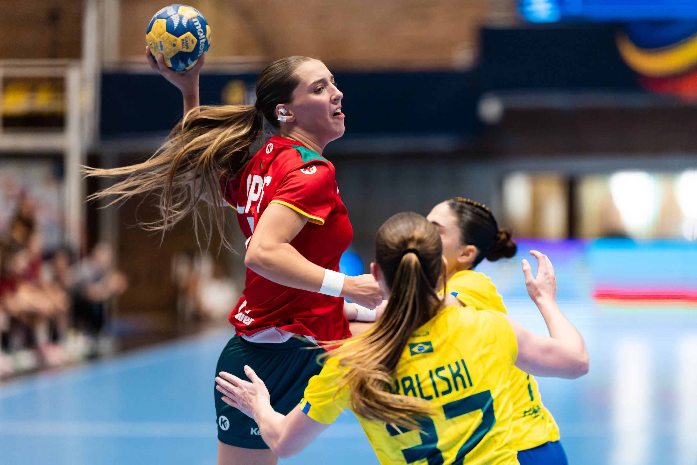 An athlete in a red jersey leaps to pass a handball while being defended by two opponents in yellow jerseys.