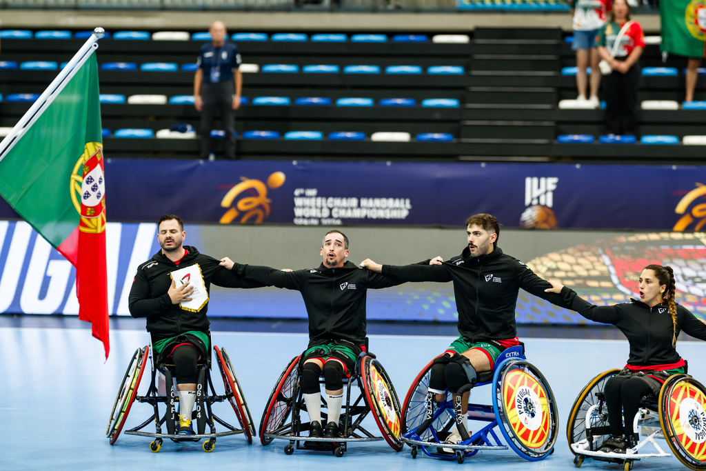 Four athletes in wheelchairs, wearing matching uniforms, pose together on an indoor sports court with a Portuguese flag and championship banners in the background.