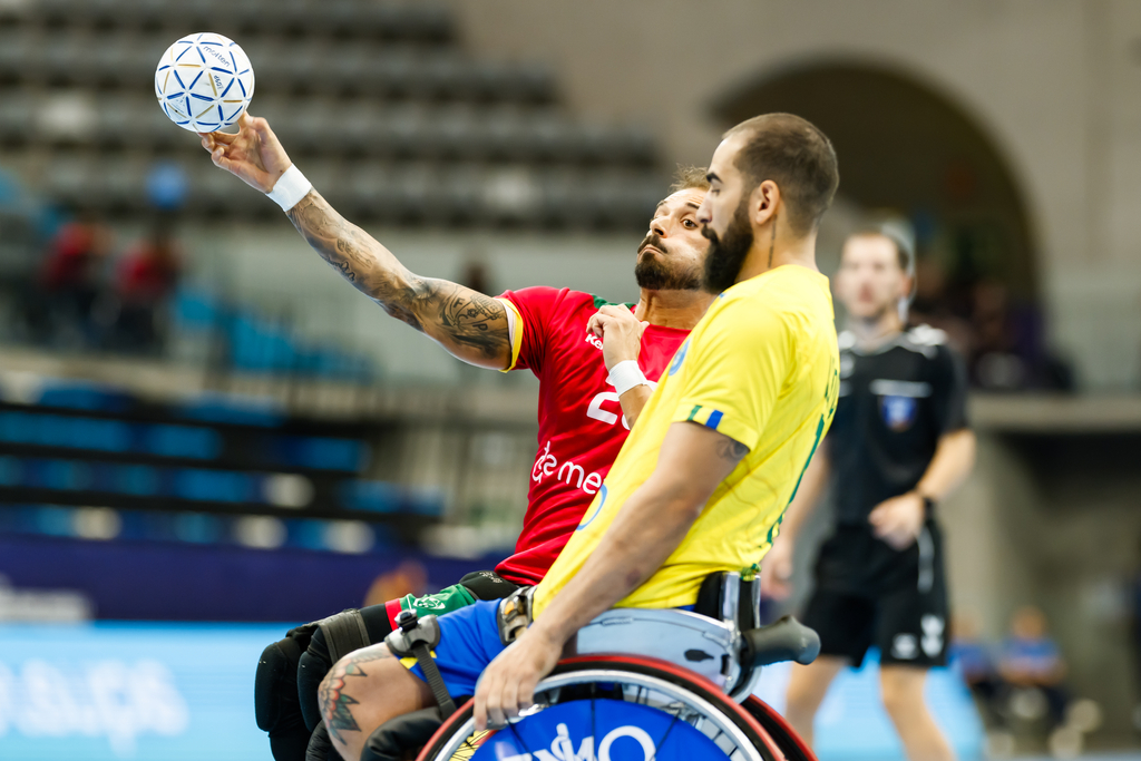 Two athletes in wheelchairs compete in a handball match, with one player in a red jersey holding the ball high while another in a yellow jersey approaches.