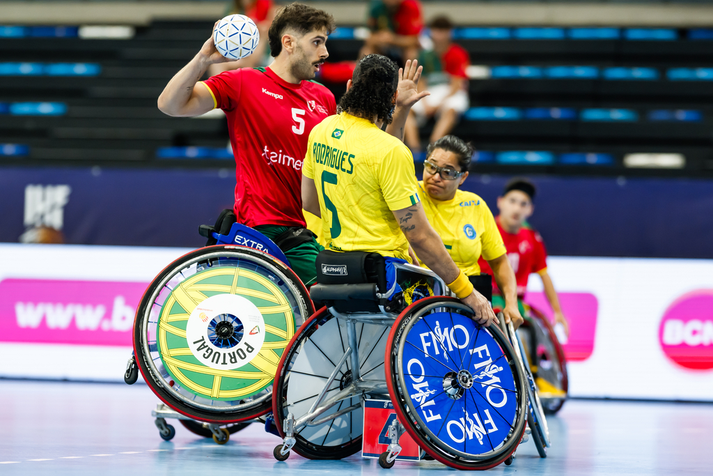 Two athletes in wheelchairs, one in a red jersey and one in a yellow jersey, are engaged in a wheelchair handball match, with the player in red holding the ball.