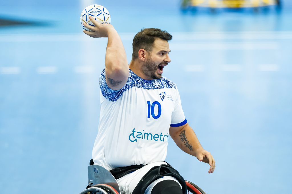 An athlete in a wheelchair, wearing a white jersey with the number 10, raises a handball above their head while shouting during a game.