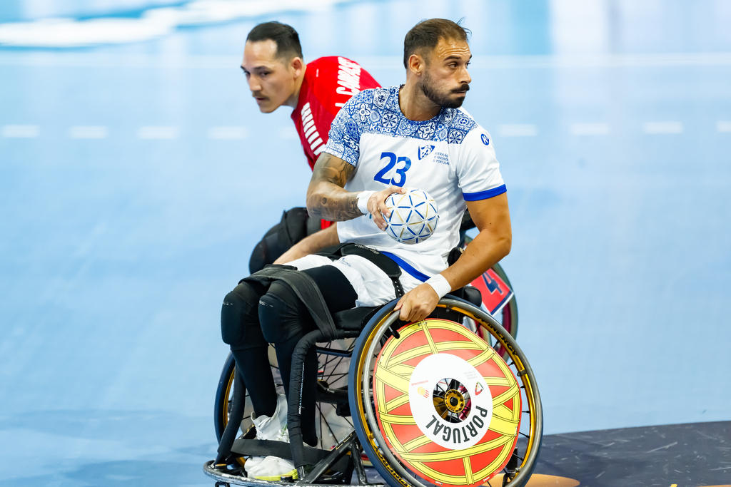 A wheelchair athlete in a white jersey with the number 23 holds a ball while another player in a red jersey trails closely behind on a blue indoor court.