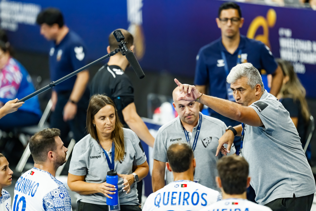 A coach gestures emphatically while speaking to players and staff during a sports event, with a microphone operator nearby.