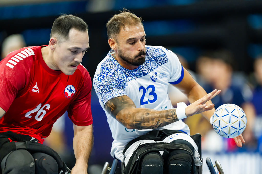 Two athletes in wheelchairs compete in a handball match, with one in a red jersey and another in a white jersey with blue patterns, both focused on the ball.