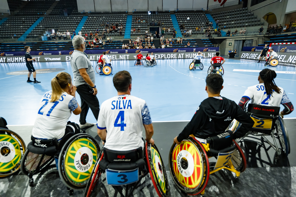 Athletes in wheelchairs compete in a seated sports event within an indoor arena, with spectators watching from the stands.