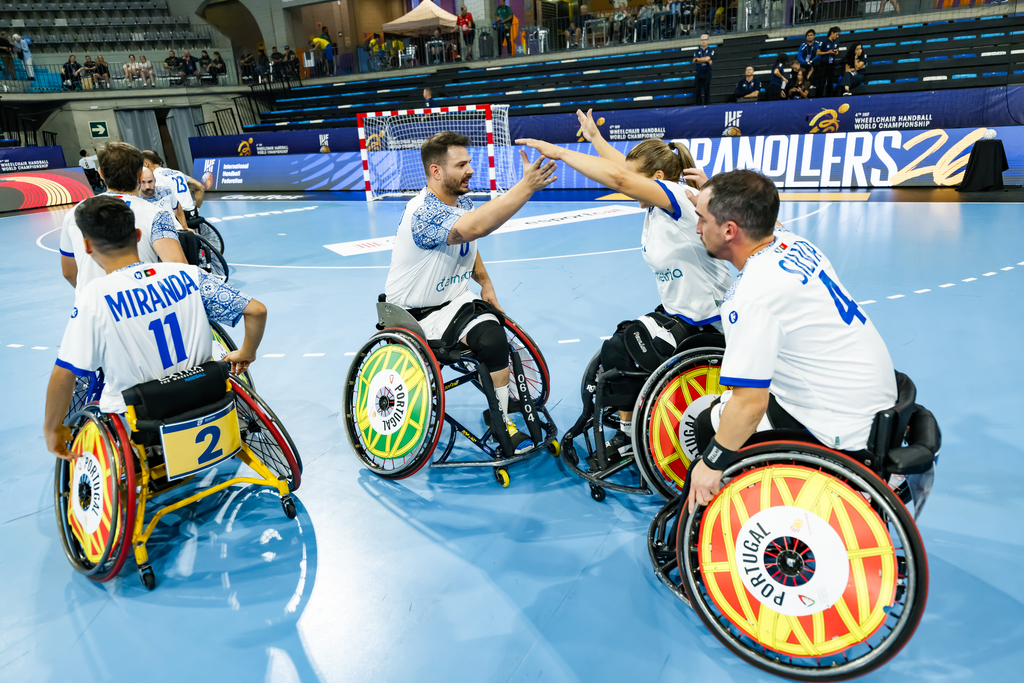 A group of wheelchair athletes in white jerseys celebrate on a blue indoor court during a wheelchair handball match.