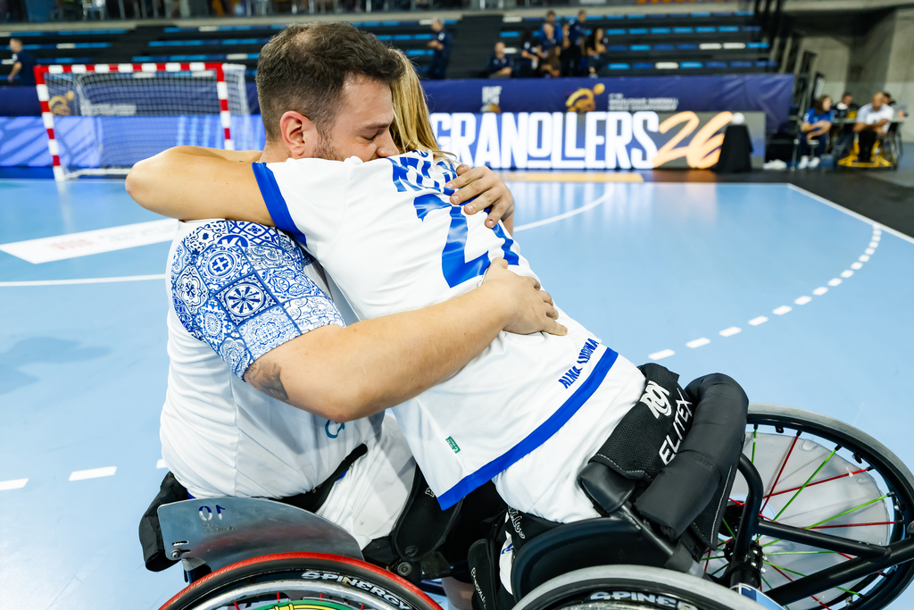 Two athletes in wheelchairs embrace warmly on a blue indoor sports court, with a “Granollers26” banner visible in the background.
