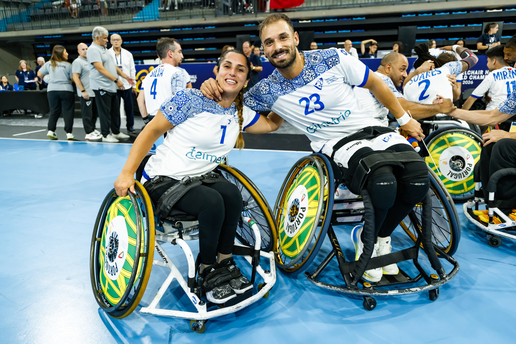 Two athletes in wheelchairs, wearing matching jerseys with numbers 1 and 23, smile together on a blue court during a wheelchair handball event.