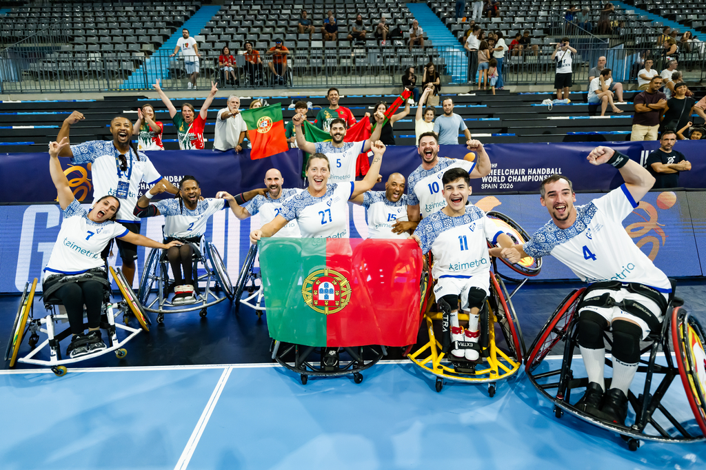 A group of wheelchair handball players in Portugal's national team uniforms celebrate on the court with their flag, surrounded by cheering fans at the 2022 Wheelchair Handball World Championship in Spain.