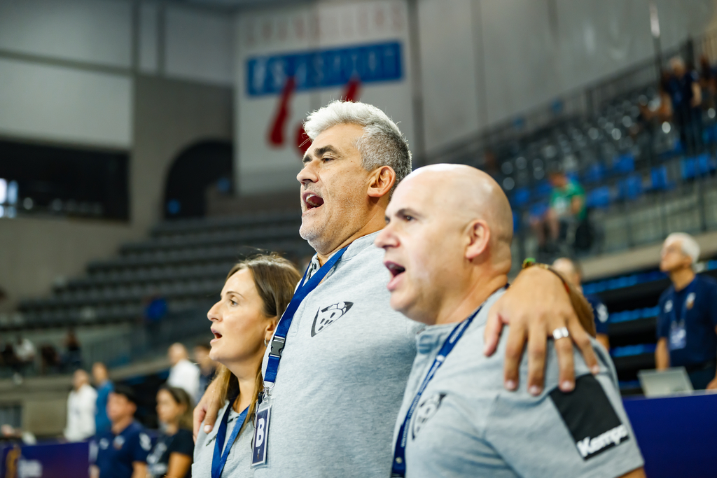 Three individuals in matching gray shirts and lanyards are intently watching an event, with one placing a hand on another's shoulder as they shout in unison.