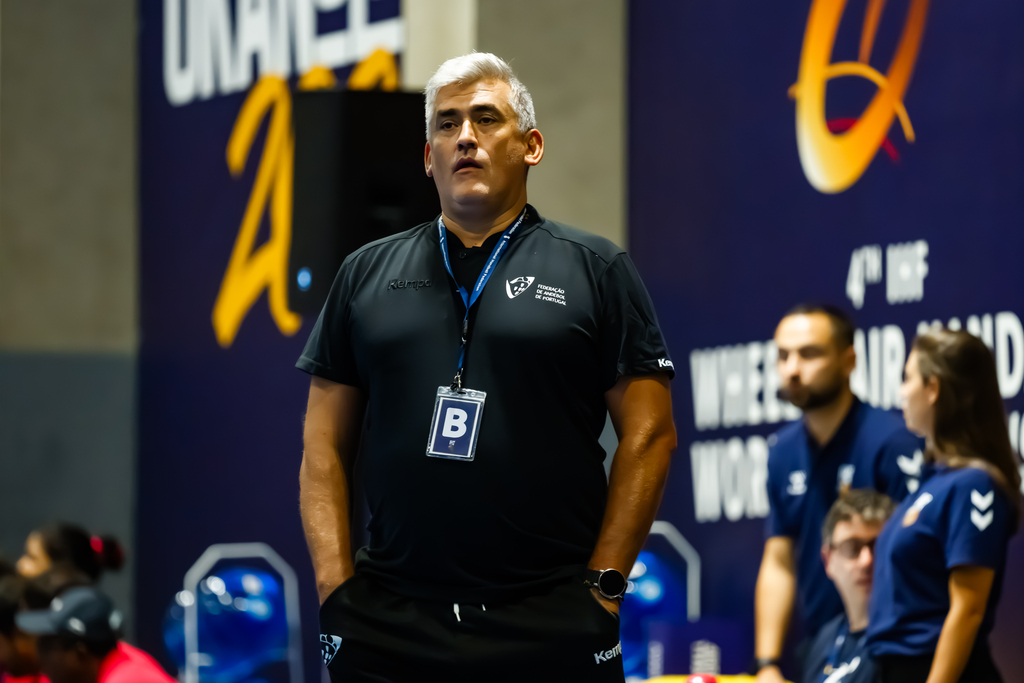 A coach in a black uniform stands attentively during a wheelchair rugby match, with event branding visible in the background.