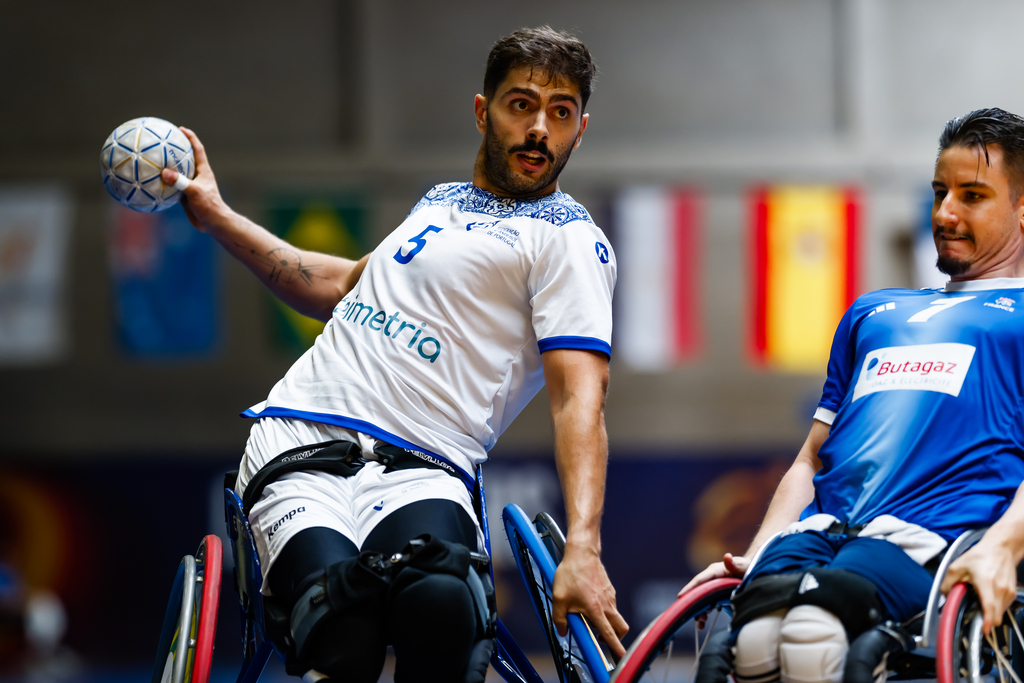 Two athletes in wheelchairs compete in a handball match, with one player in a white jersey preparing to throw the ball.