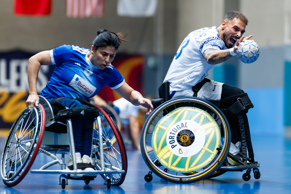 Two athletes in wheelchairs compete in a fast-paced indoor sport, one in blue and one in white, with the white-clad player holding the ball.