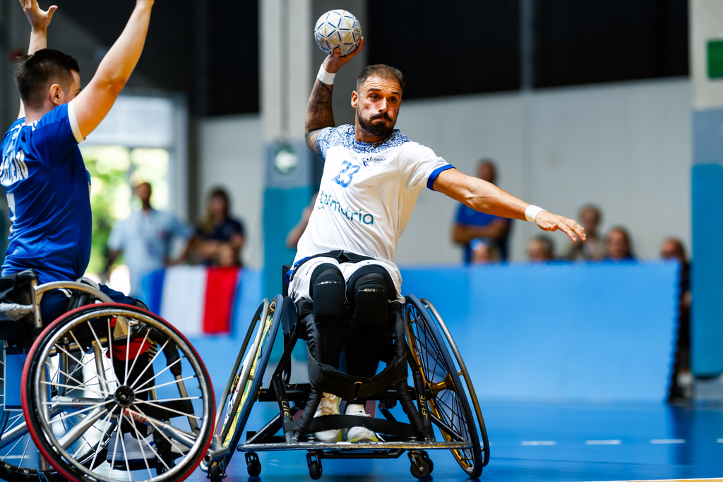 An athlete in a wheelchair prepares to throw a ball during a wheelchair handball game, while an opponent in a blue jersey defends.