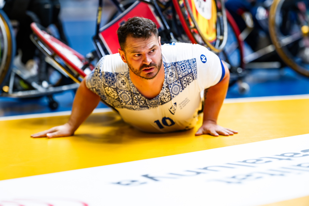 An athlete in a patterned jersey with the number 10 is on their hands and knees on a yellow court, with wheelchairs visible in the background.
