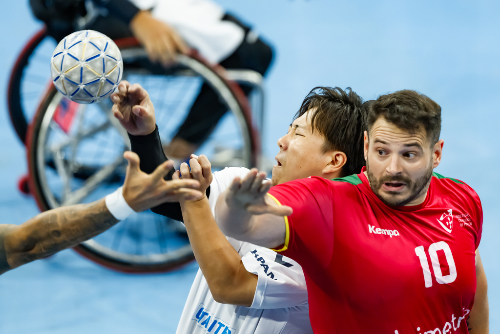 Two athletes compete for a handball during a wheelchair handball match, with one player in a red jersey and another in a white jersey reaching for the ball.