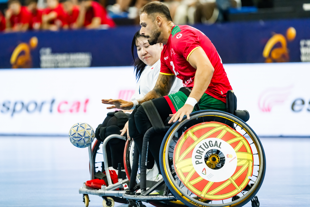 Two athletes in wheelchairs compete in a wheelchair rugby match, with one player in a red jersey and the other in white, as the ball rolls near their wheels.