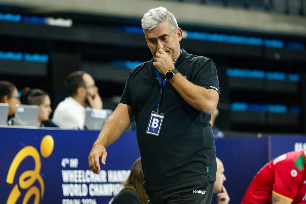 A coach in a black uniform with a "B" badge covers his mouth while walking during a wheelchair handball event.