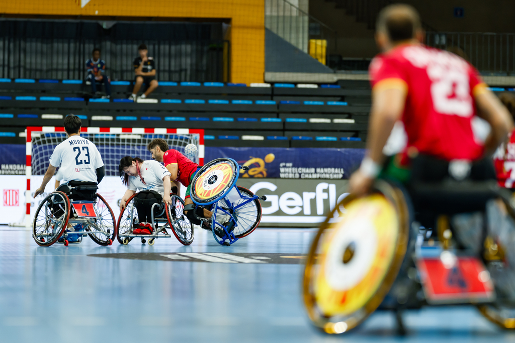 Athletes in wheelchairs compete in an indoor wheelchair handball match, with one player preparing to shoot toward the goal.